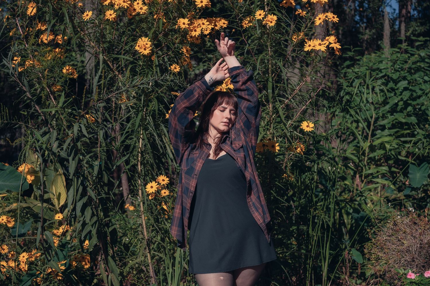 Woman in plaid shirt poses gracefully among vibrant yellow wildflowers in a lush garden setting.