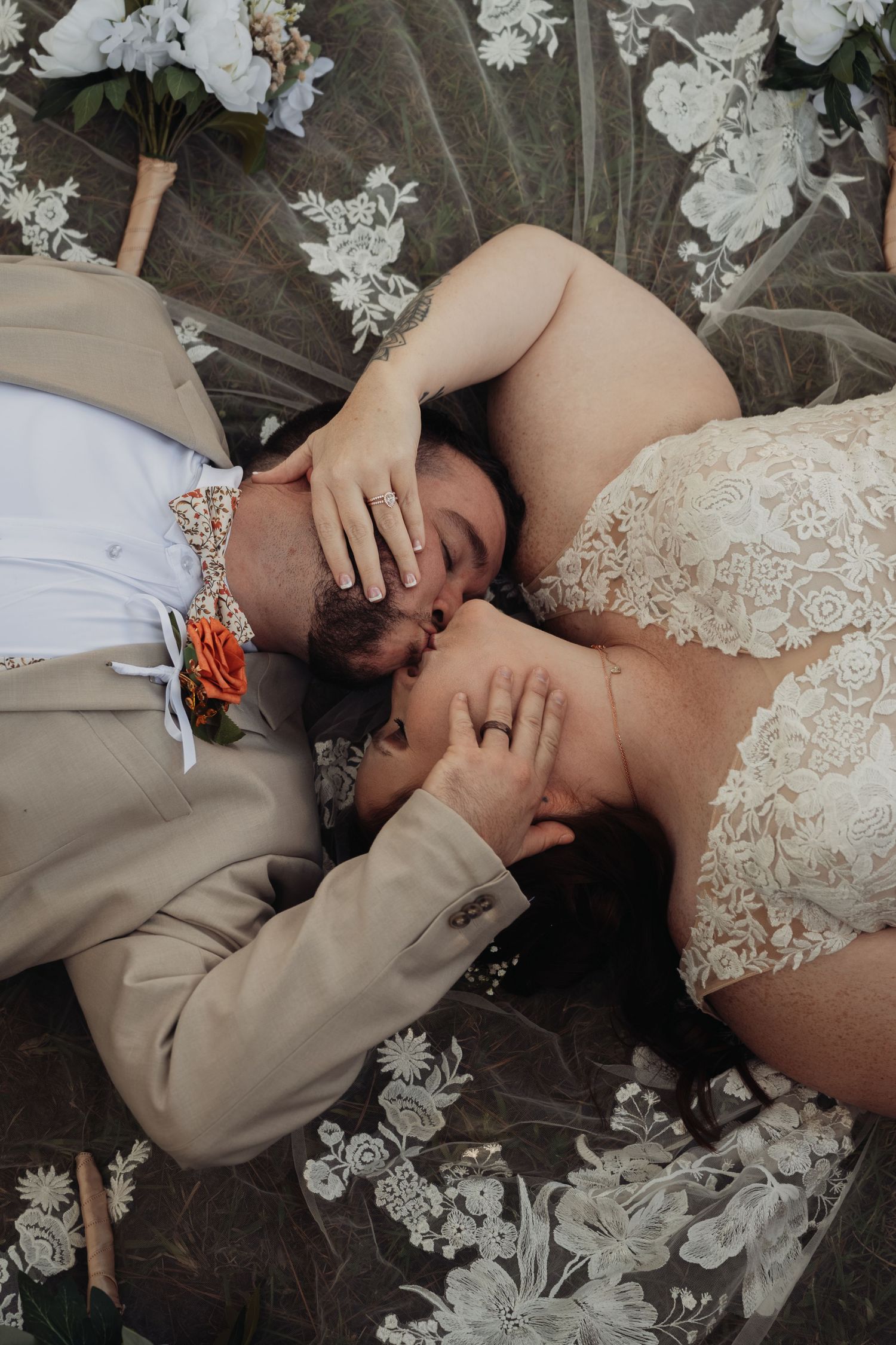 Overhead view of a bride and groom lying on lace-covered ground, sharing a kiss with a bouquet nearby.