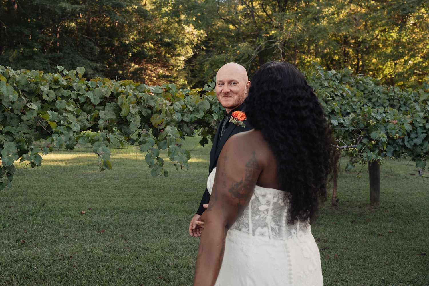 Bride in strapless lace gown glances back at her groom standing among green vineyard vines.