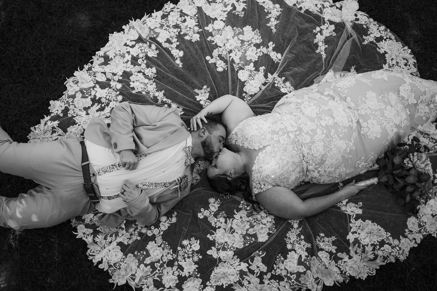 Black and white overhead photo of a bride and groom lying face to face on a wide lace wedding veil spread out like a floral circle.