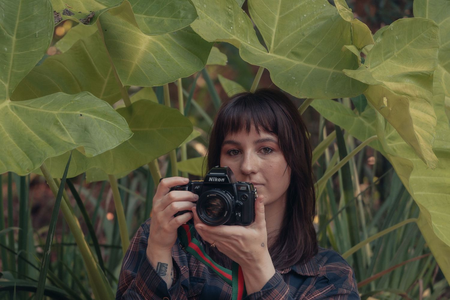 A tattooed woman with bangs holds a camera up to photograph amid large tropical green leaves outdoors.