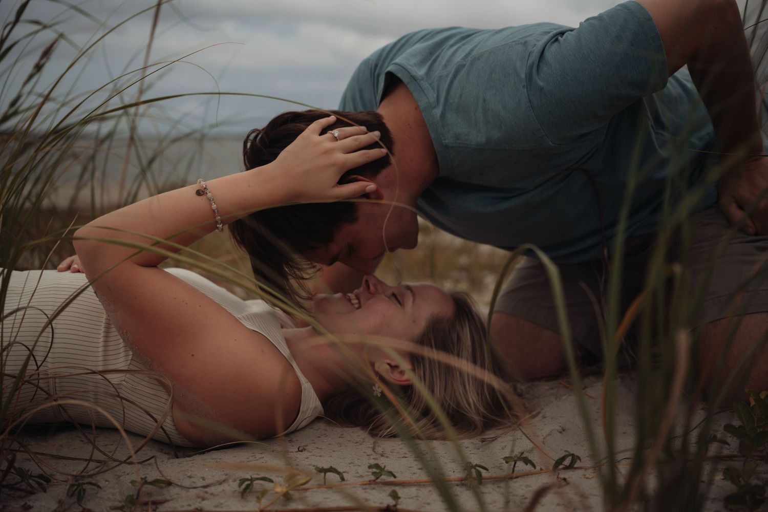 A couple shares a tender kiss on a sandy beach, surrounded by tall grass, as the woman lies down and the man leans over her.