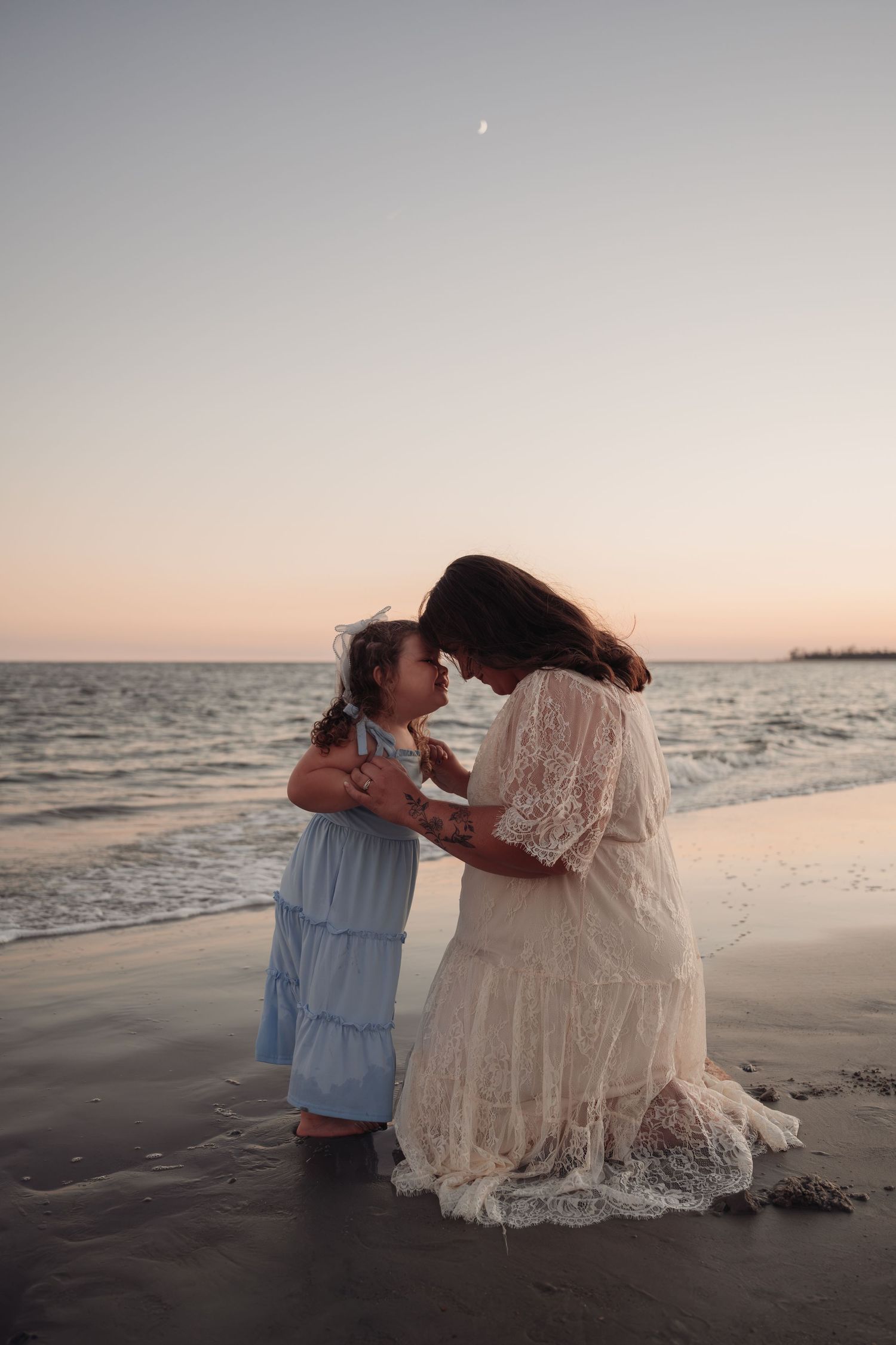 Mother in a cream lace dress and young daughter in a blue dress touch foreheads on a sandy beach at sunset with a crescent moon above.