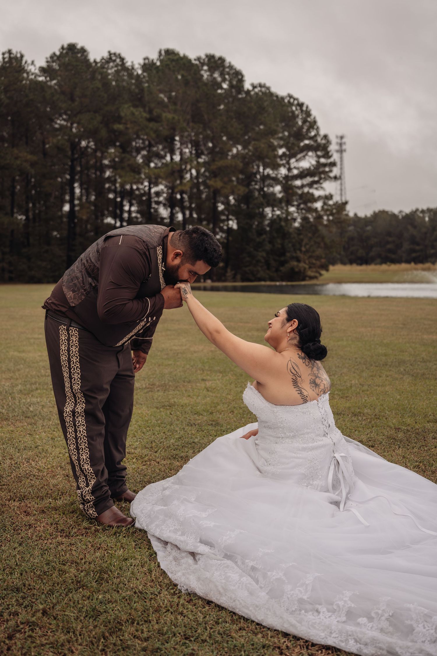 Groom in brown suit kisses bride's hand as she sits in white gown on grass near a lake surrounded by trees.