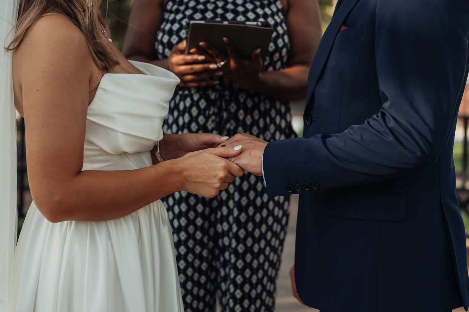 Bride and groom exchange rings during an outdoor wedding ceremony with an officiant present.