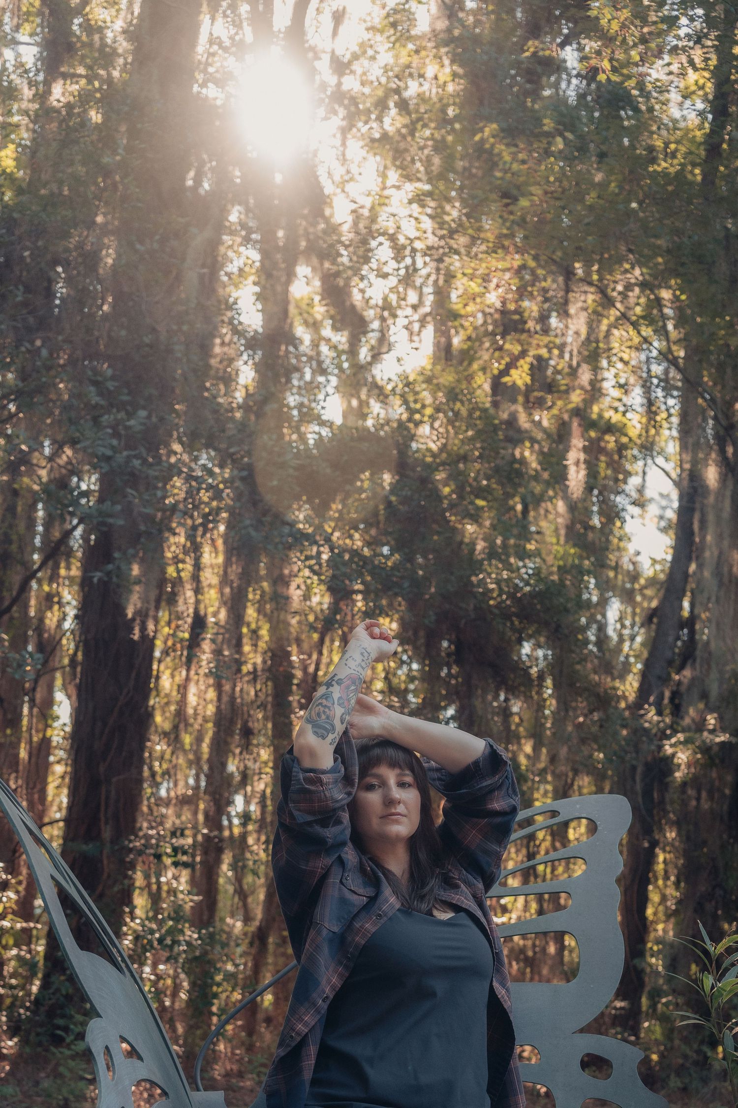 Woman with tattoos poses in sunlit forest with butterfly sculpture, wearing plaid shirt, arms raised overhead.