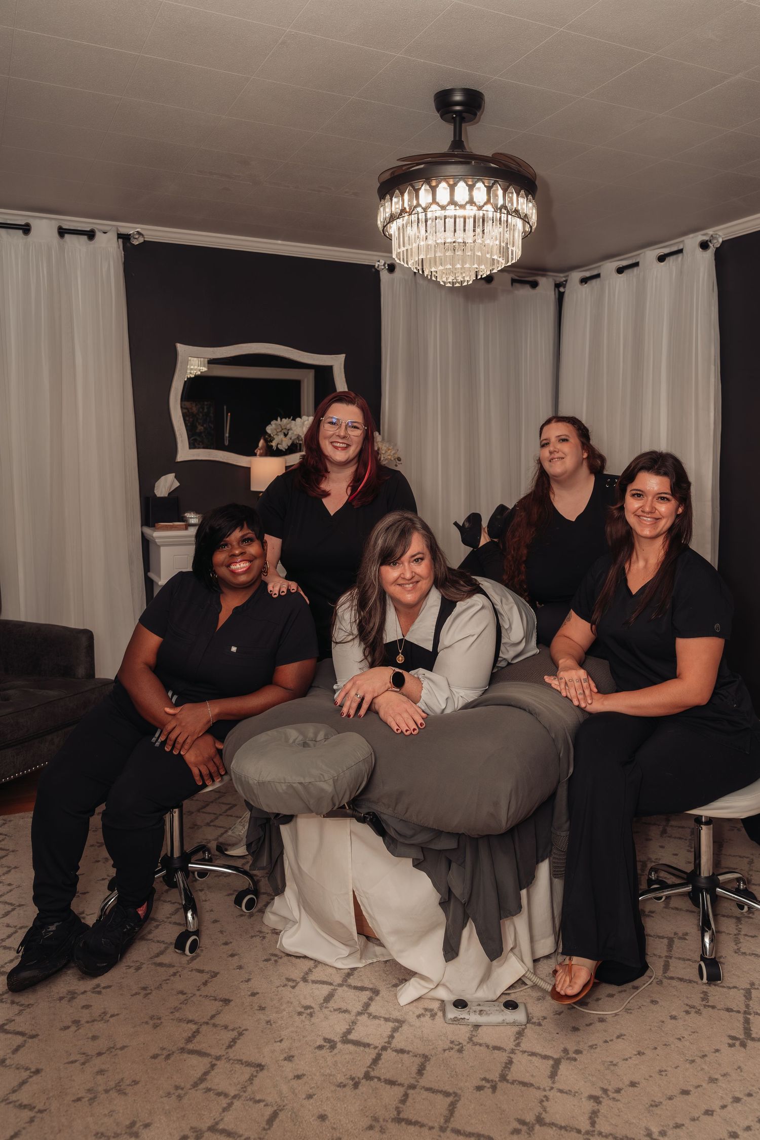 Five smiling women in black uniforms pose around a massage table with a crystal chandelier in an elegant spa treatment room.