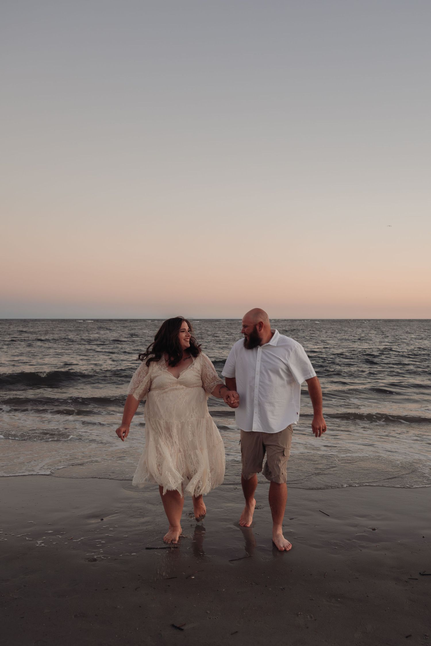 Couple holding hands and smiling at each other while walking barefoot along the ocean shore at sunset during a beach engagement session.