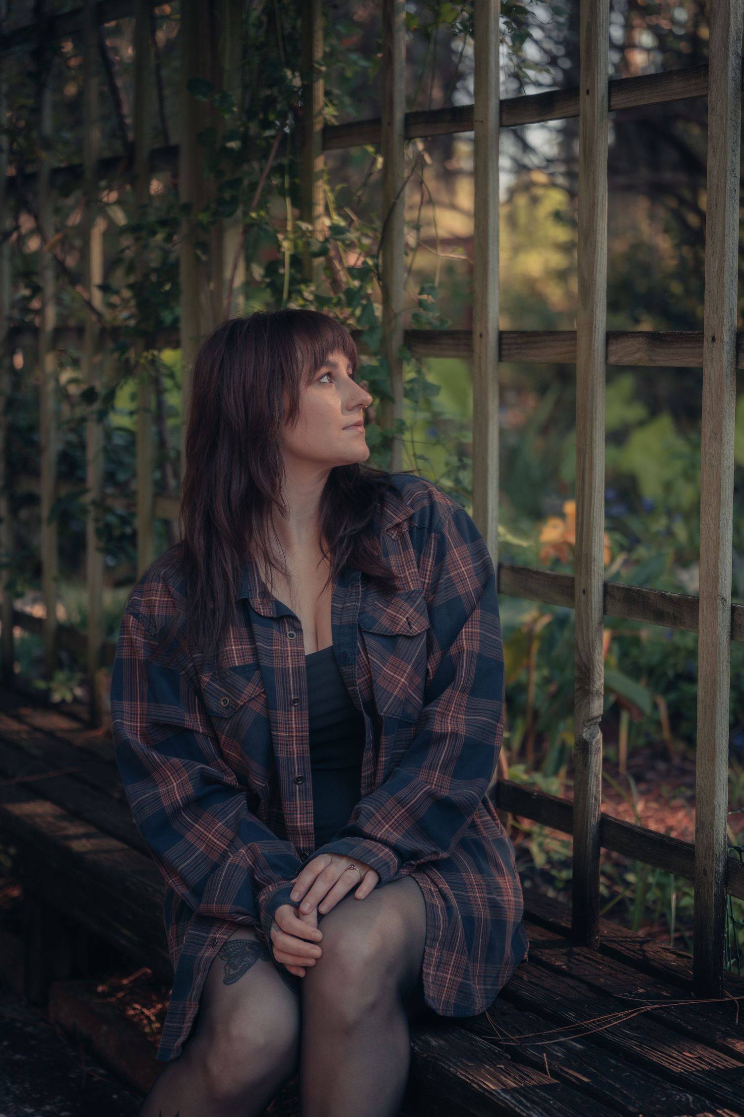 Young woman in plaid shirt sits pensively on wooden bench in rustic garden greenhouse setting.