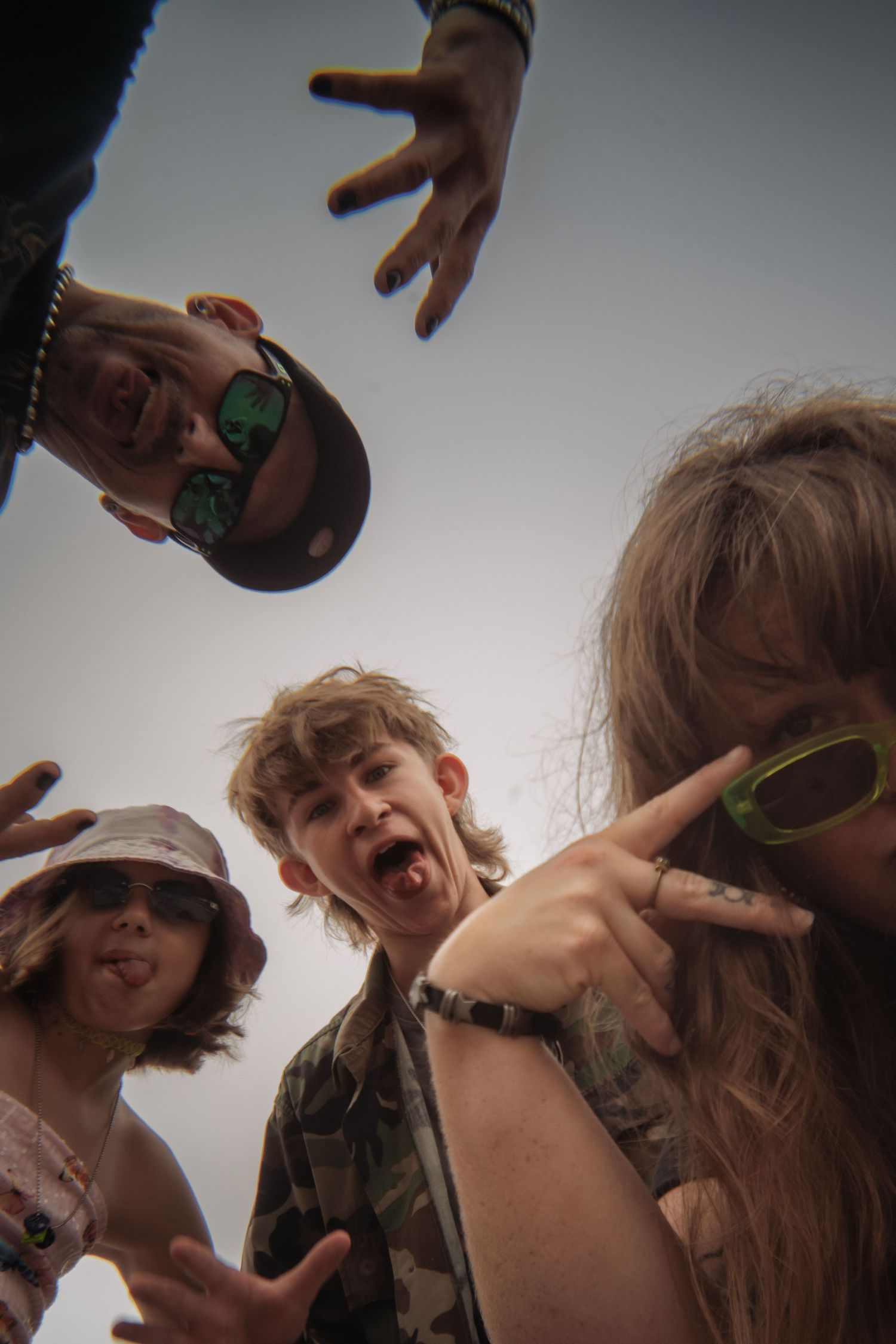 Four playful friends in sunglasses and hats lean over the camera in a low-angle shot, sticking out tongues and flashing hand gestures.