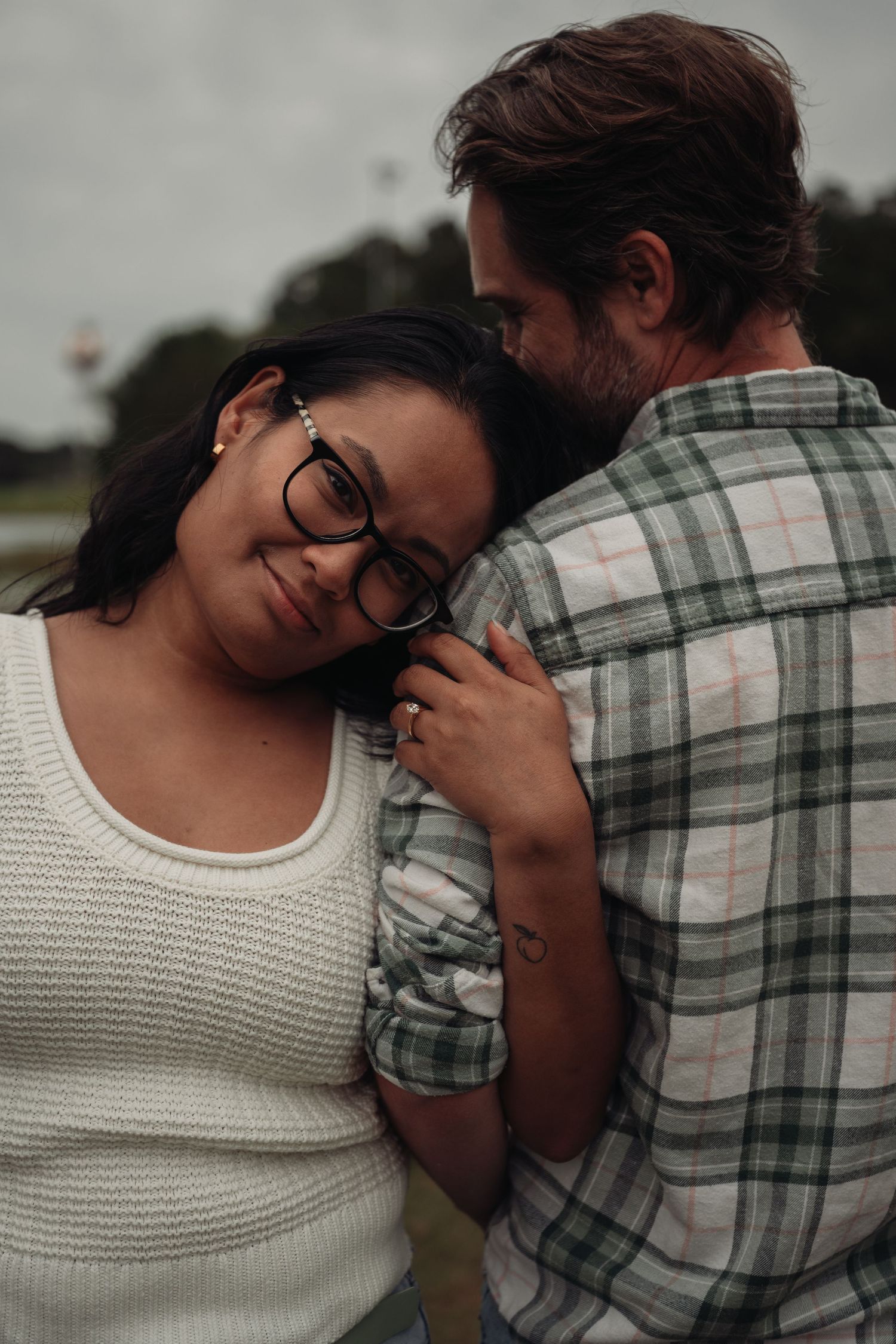 A woman with glasses rests her head on a man's chest outdoors, sharing a tender embrace on a cloudy day.