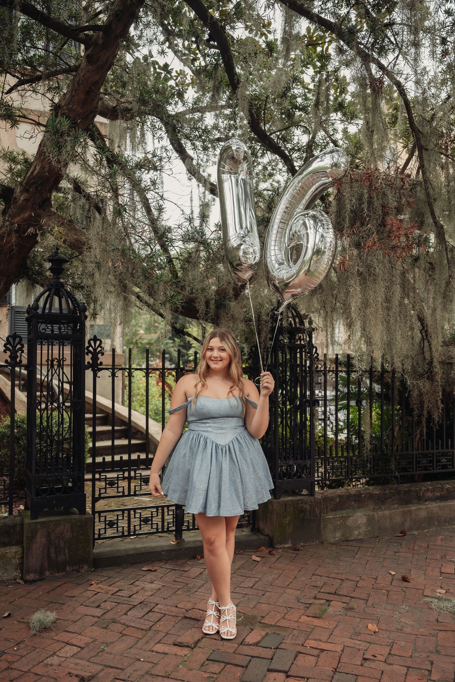 A teen girl in a blue dress holds silver '16' balloons on a brick path with Spanish moss trees and iron fence.