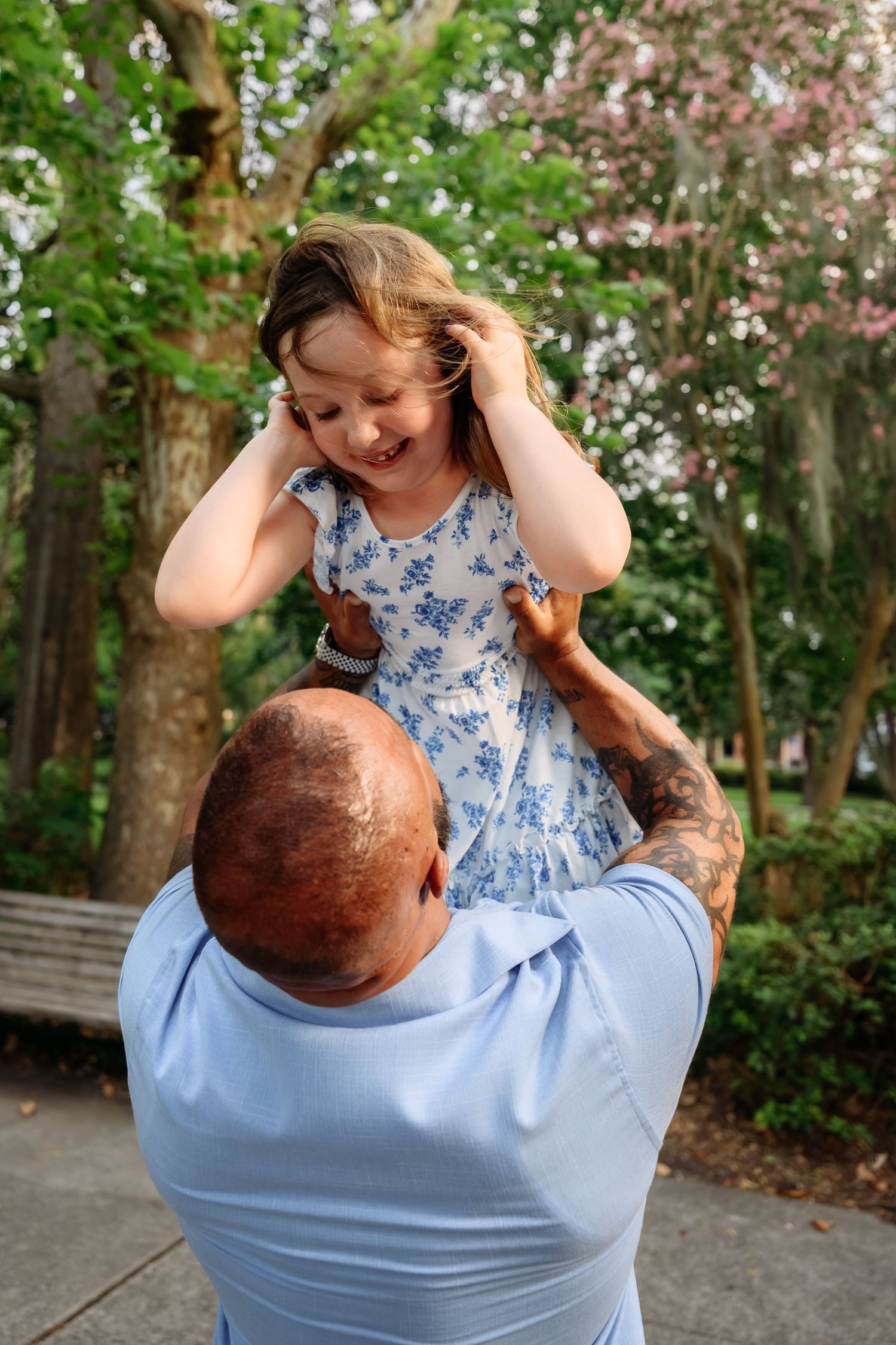 A bald father lifts his laughing young daughter in a blue floral dress outdoors in a lush green park.