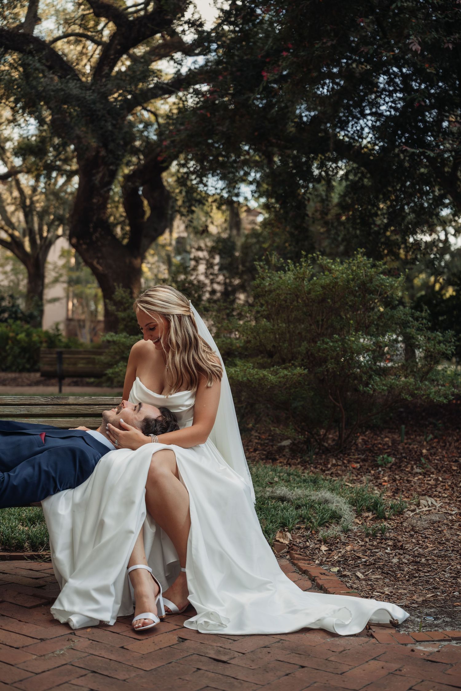 Bride in white gown sits with groom on brick path beneath oak trees in a lush park.