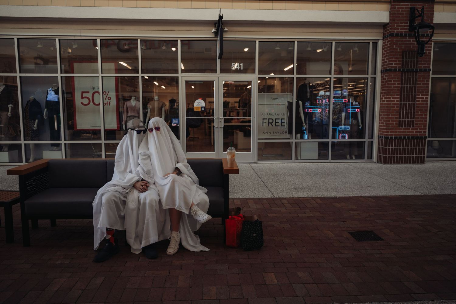 Two people dressed as ghosts in white sheets sit together on a bench outside a retail store.