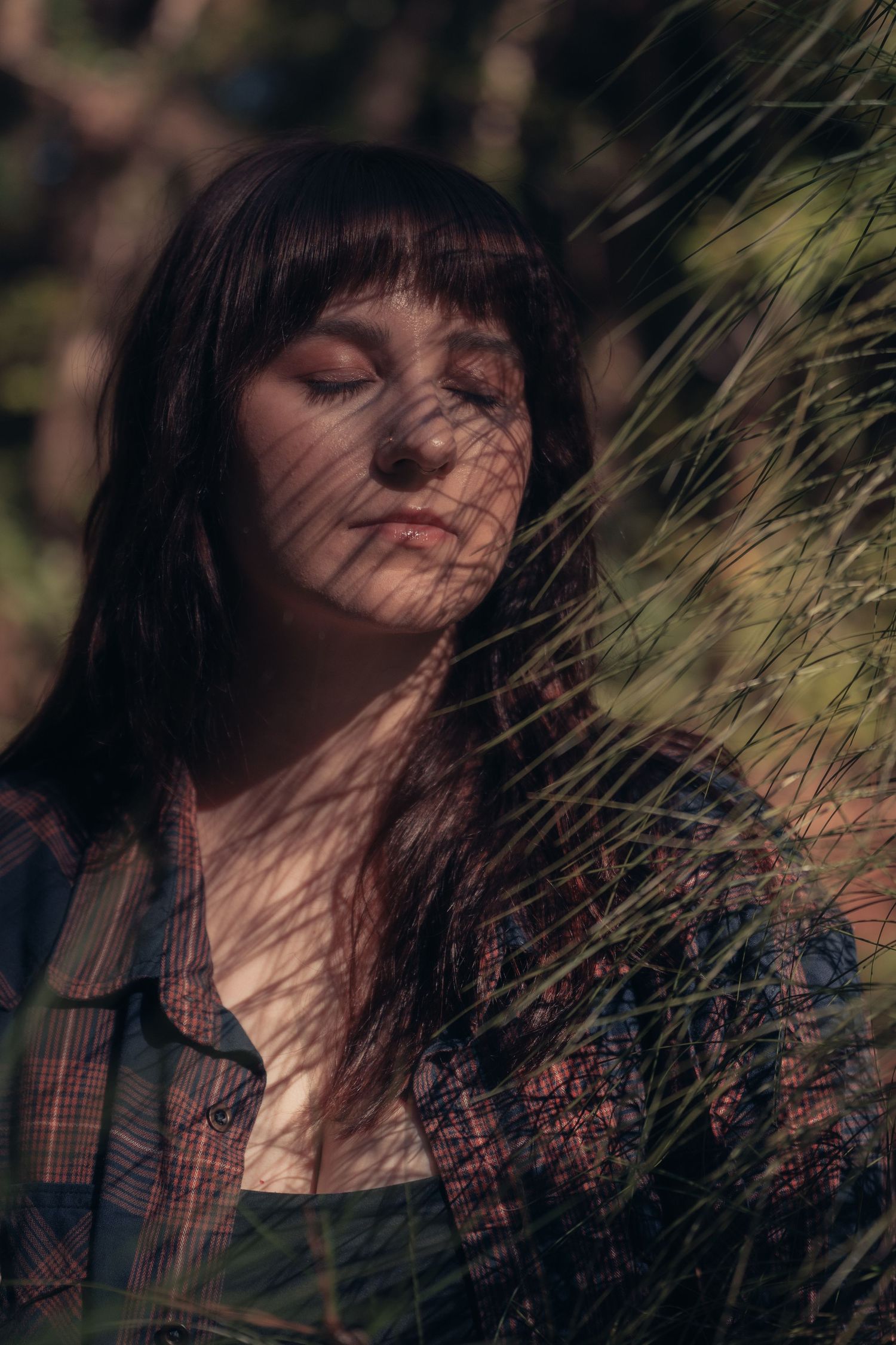Young woman with dark hair and closed eyes standing among green grass in dappled sunlight wearing a plaid shirt.