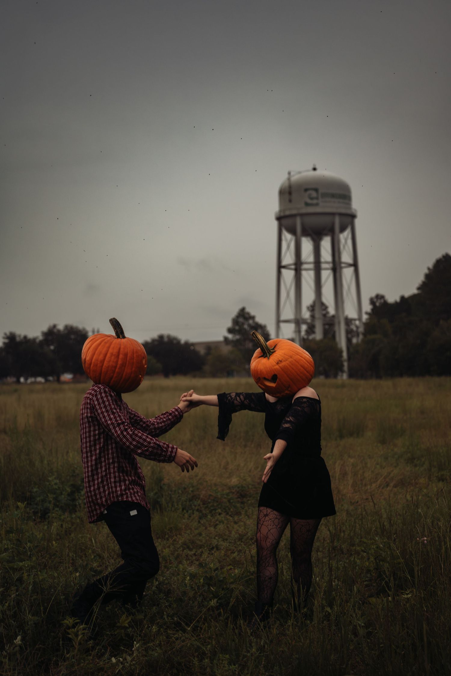 Couple with pumpkin heads dancing in a field with a water tower in the background.