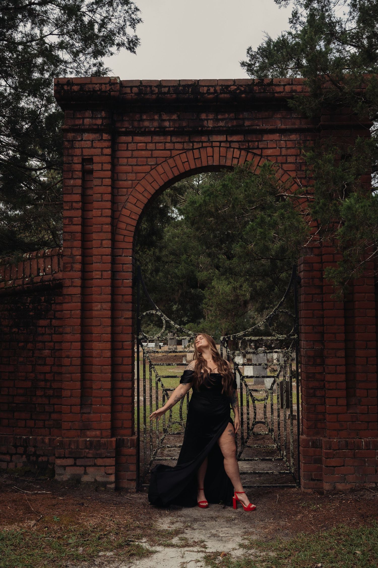 Woman in black gown and red heels poses at ornate brick archway gate surrounded by lush greenery.