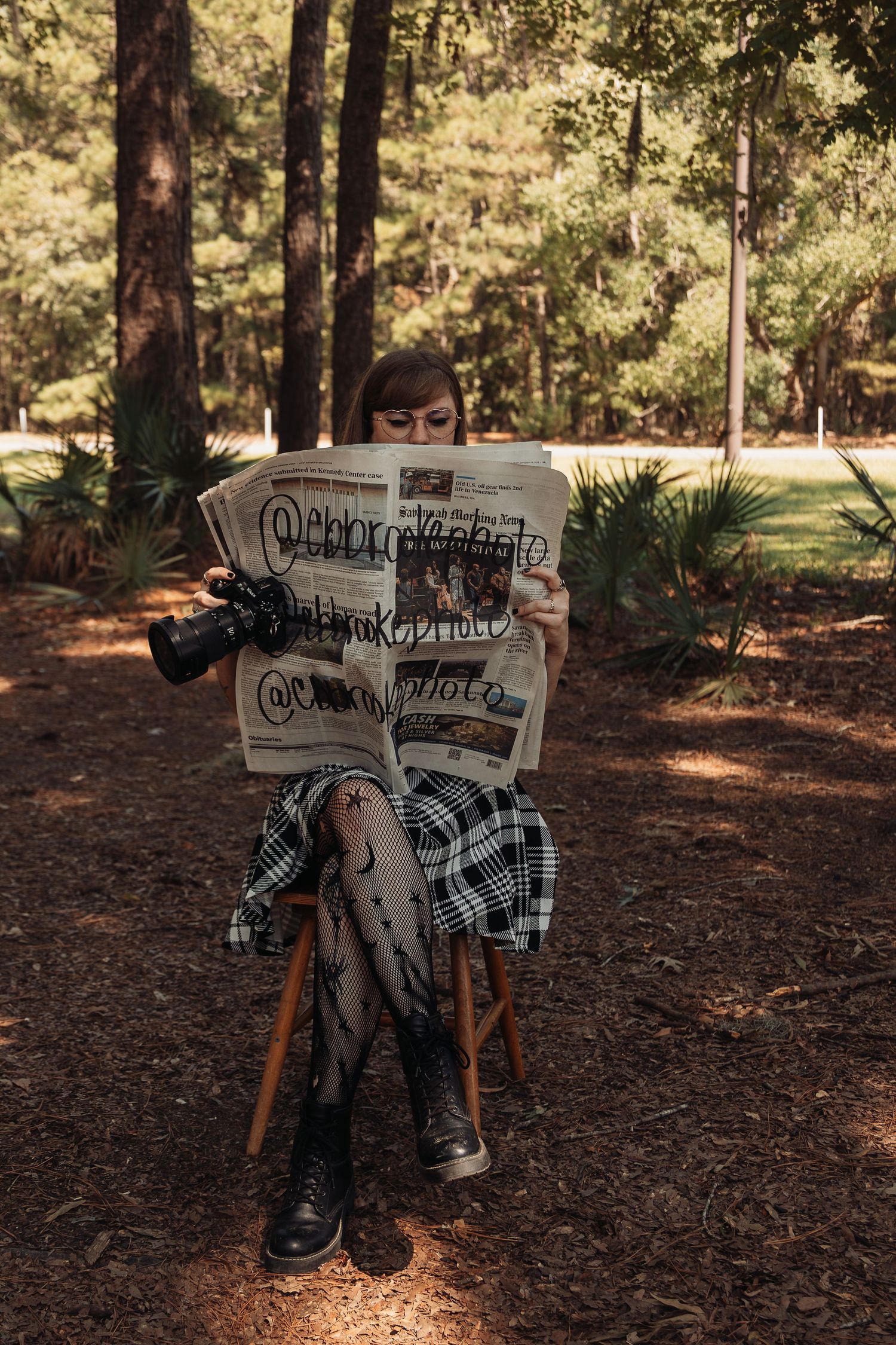 Young woman reading newspaper with camera social media handles written on it while sitting in forest.