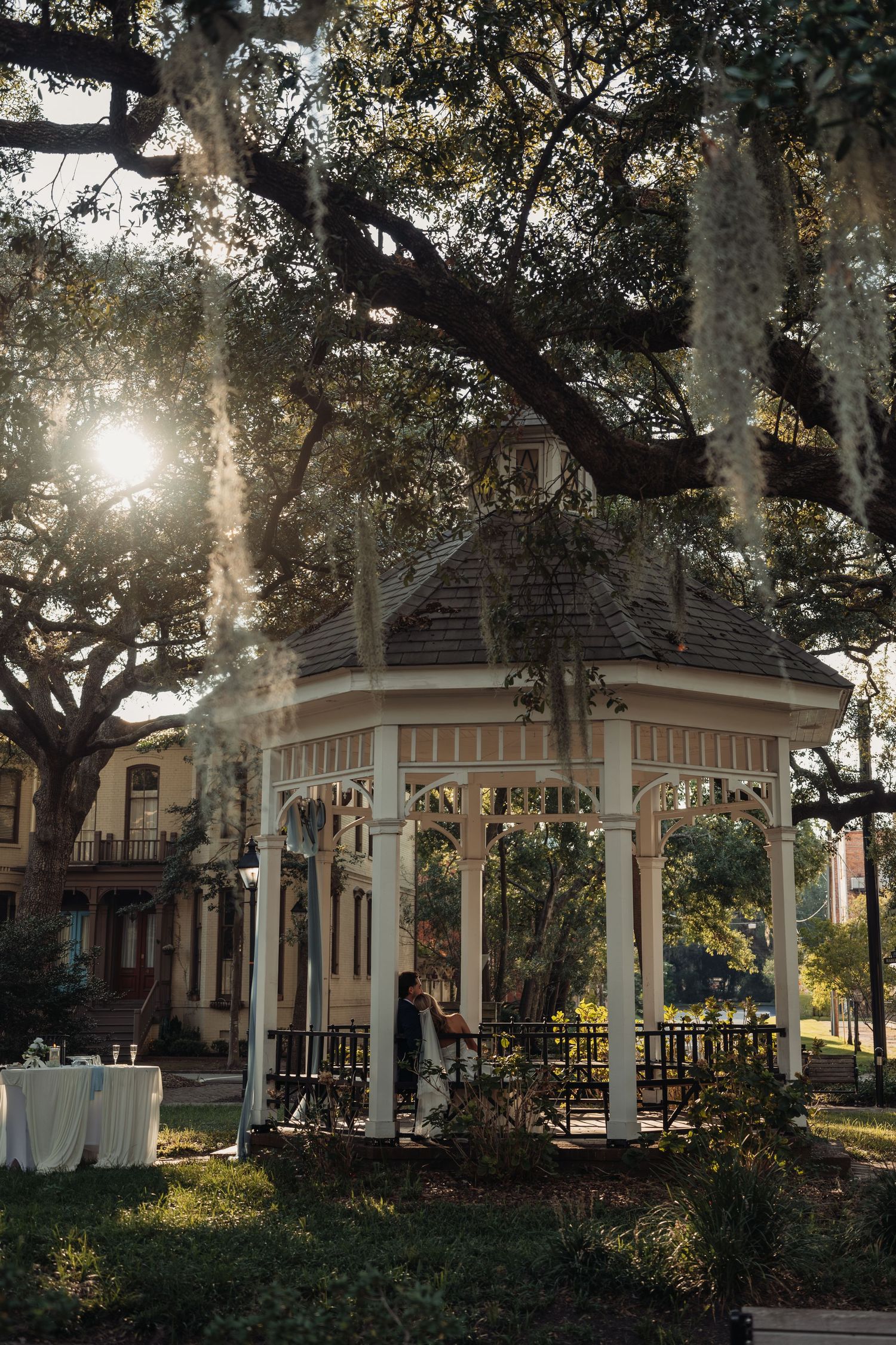 A charming white gazebo adorned with Spanish moss and sunlight in a historic park setting.