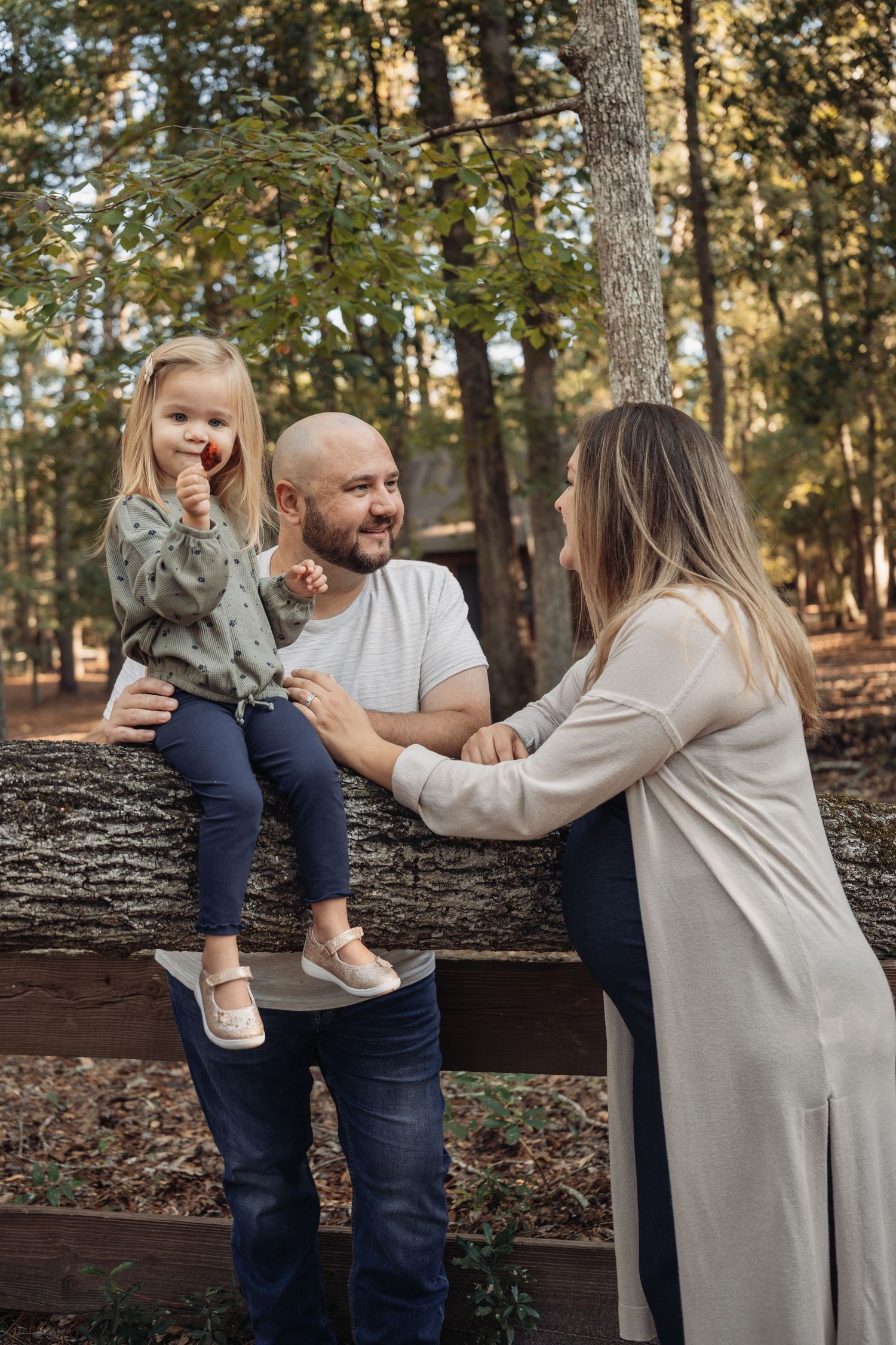 A happy family with a toddler girl sitting on a log fence in a sunny forest park setting.