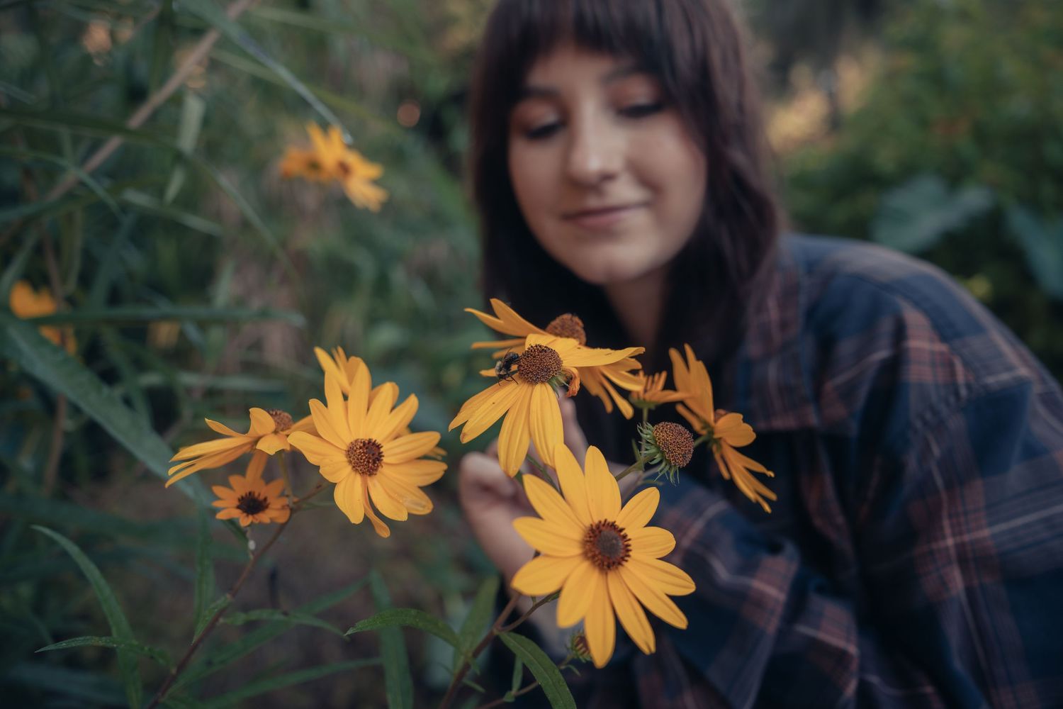 A young woman with dark hair smiles softly while holding vibrant yellow daisy flowers outdoors.