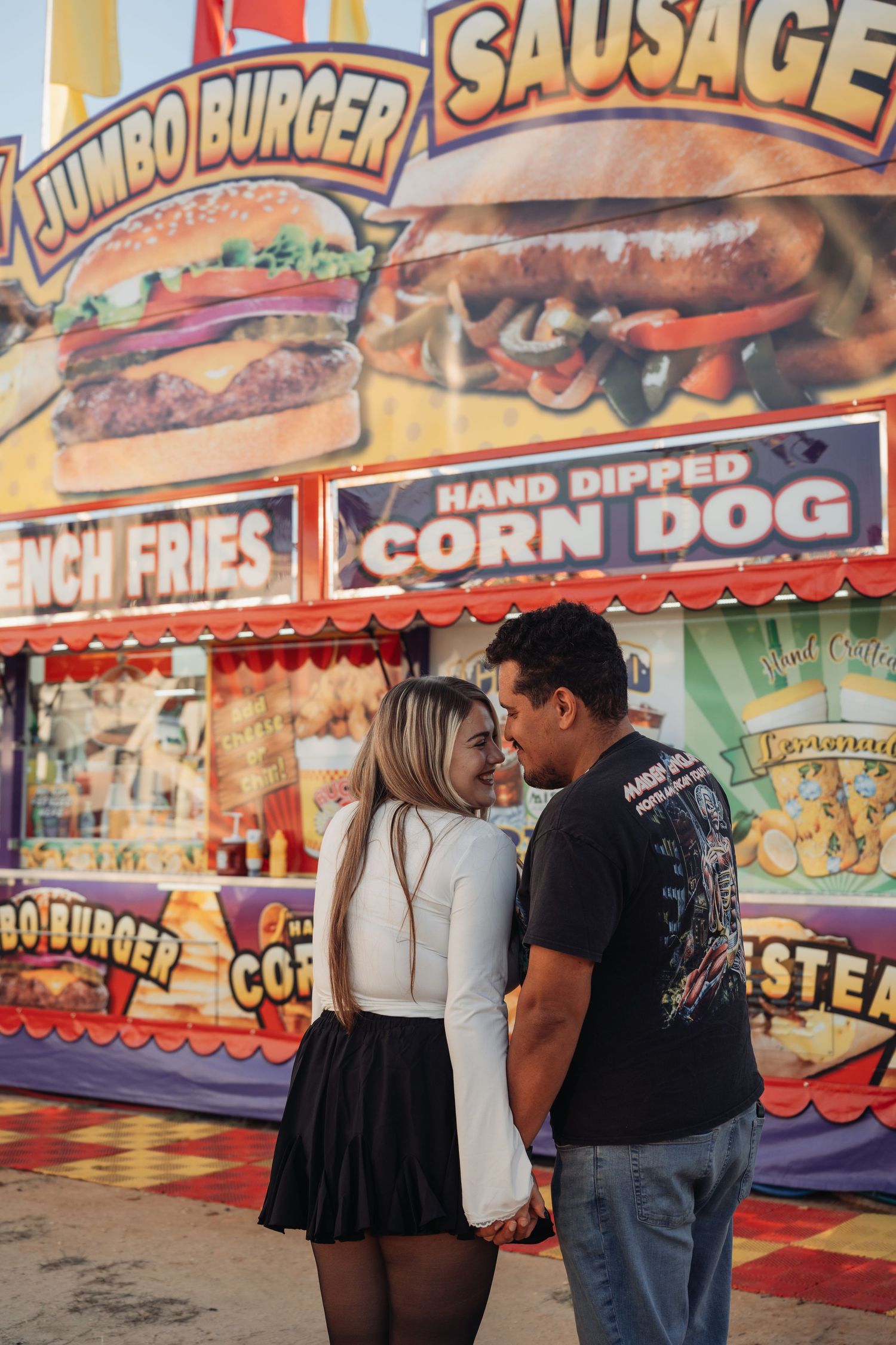 Smiling couple holding hands and gazing at each other in front of a colorful carnival food stand with corn dogs, burgers and sausage.