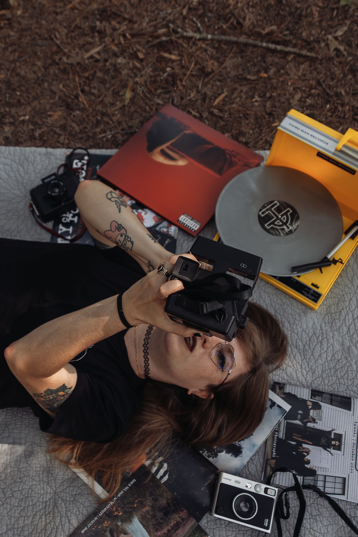 Tattooed woman lying on blanket taking photo with Polaroid camera surrounded by vinyl records outdoors.