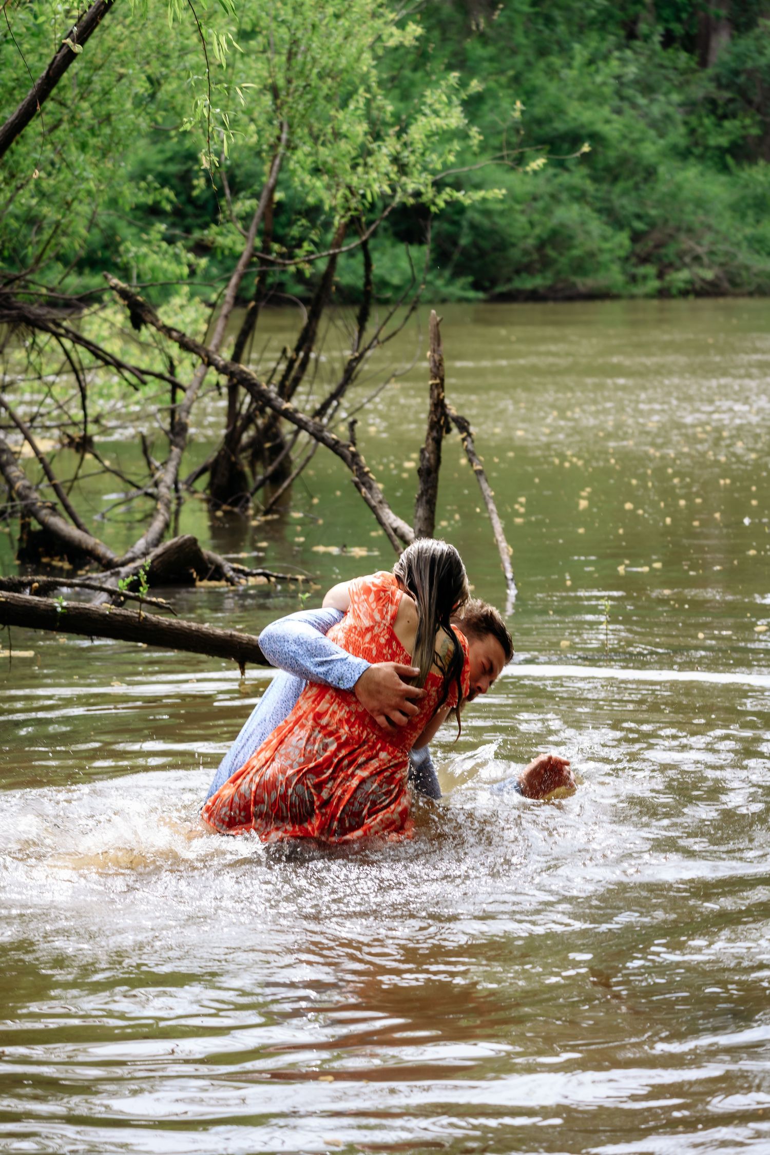 Couple splashing and embracing playfully in a river surrounded by lush green trees.