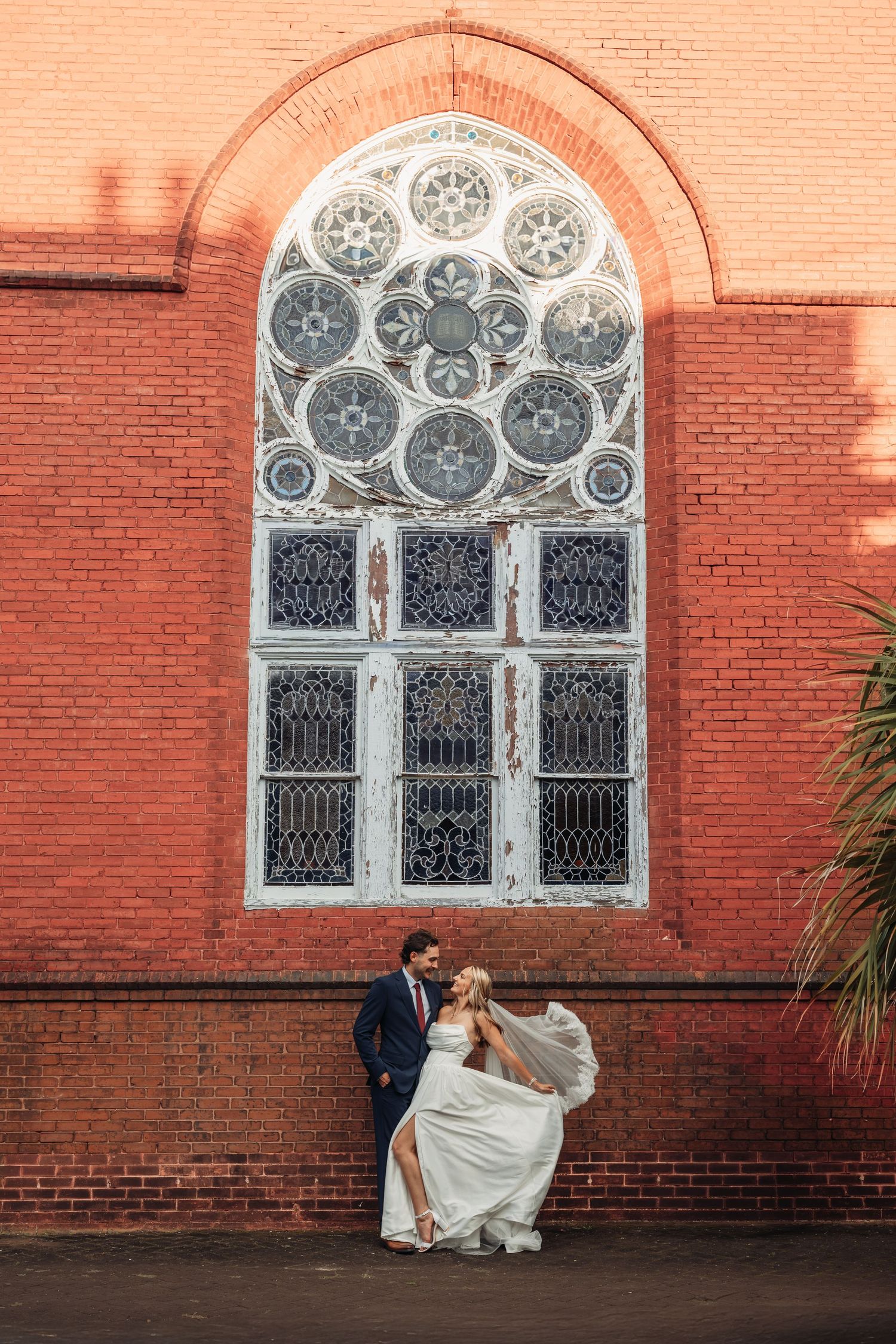 Bride and groom share a romantic moment before a stunning arched stained glass window on red brick.