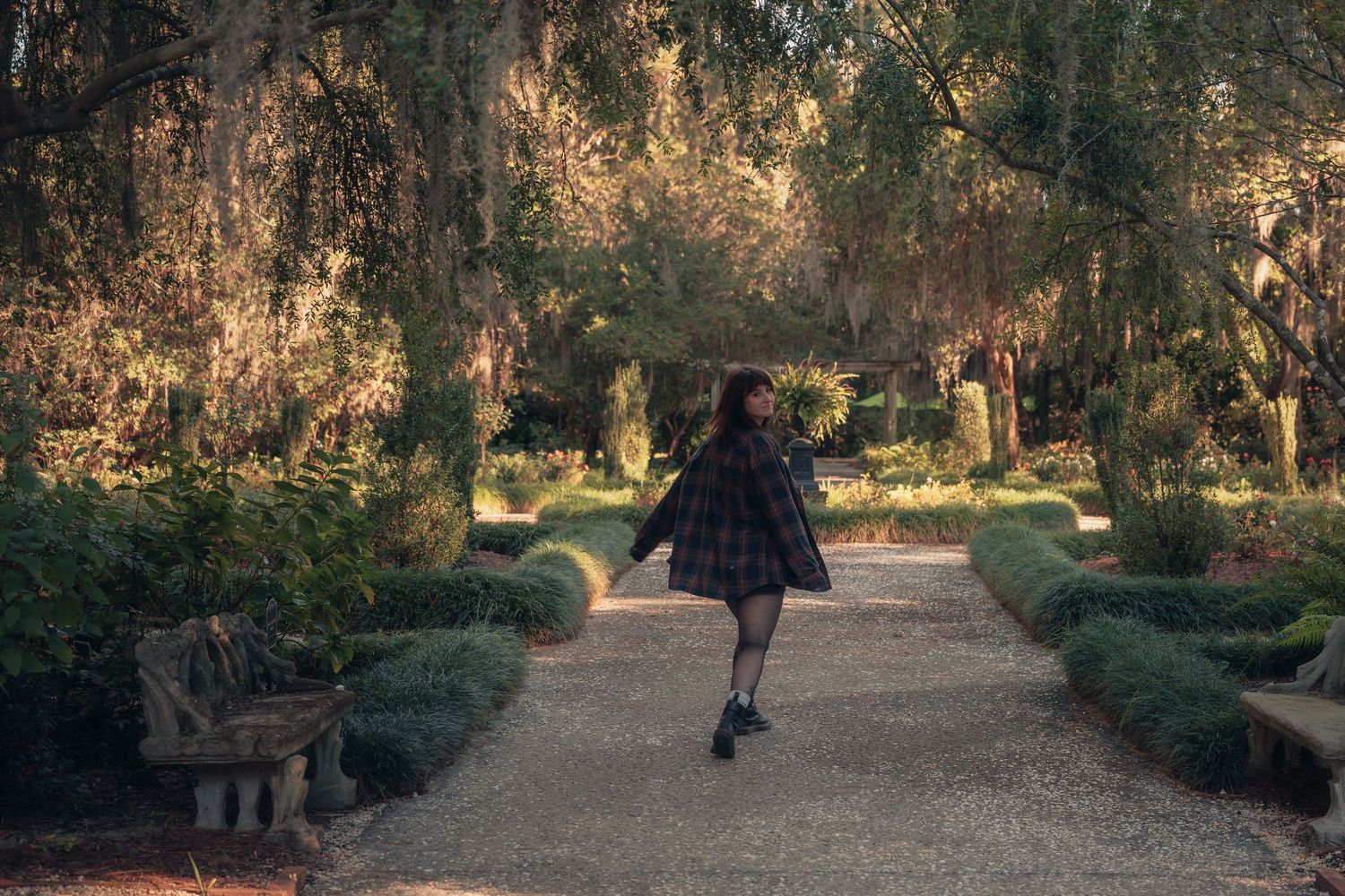 A young girl in a plaid dress walks along a garden path surrounded by lush trees and manicured hedges.