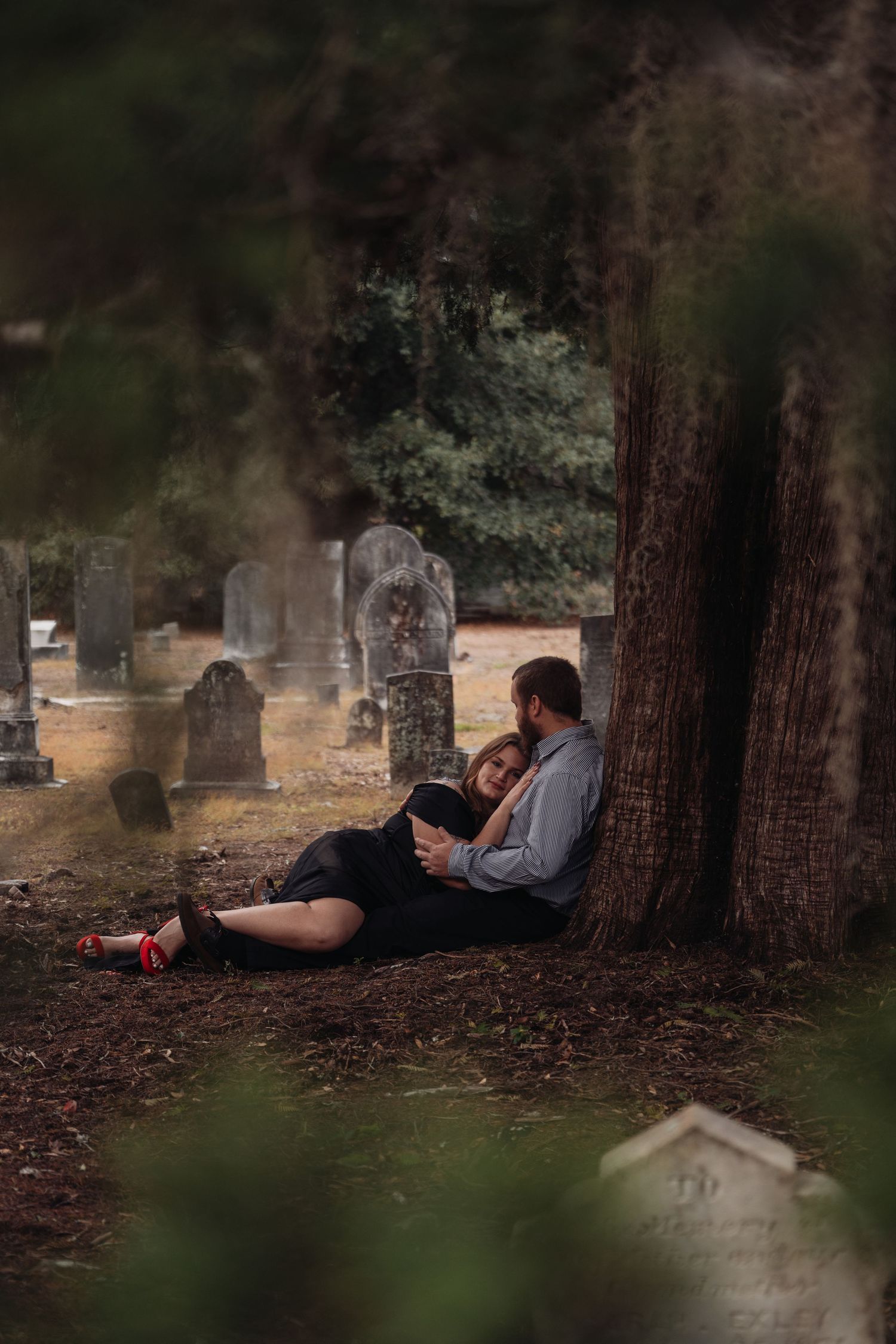 Couple sitting together against a tree in a historic cemetery, reading a book surrounded by old gravestones.