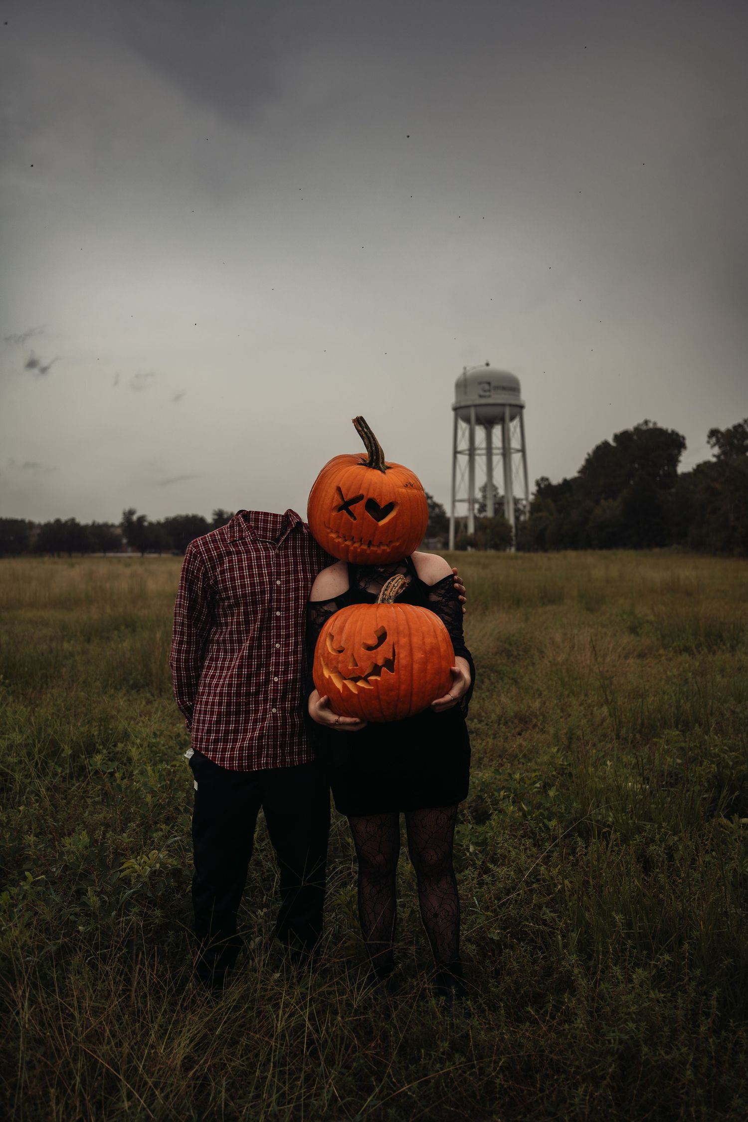 Couple holding jack-o-lanterns with pumpkin heads in a field near a water tower on a spooky Halloween day.