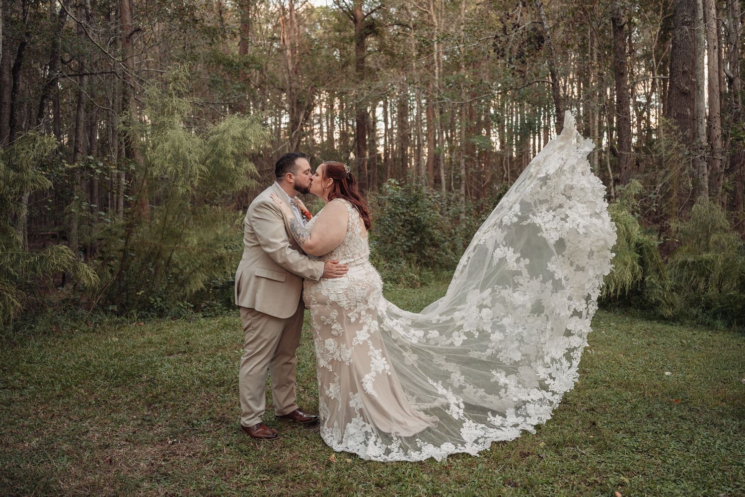 Bride and groom share a kiss in a sunlit pine forest, her long lace wedding veil flowing across the grass behind them.