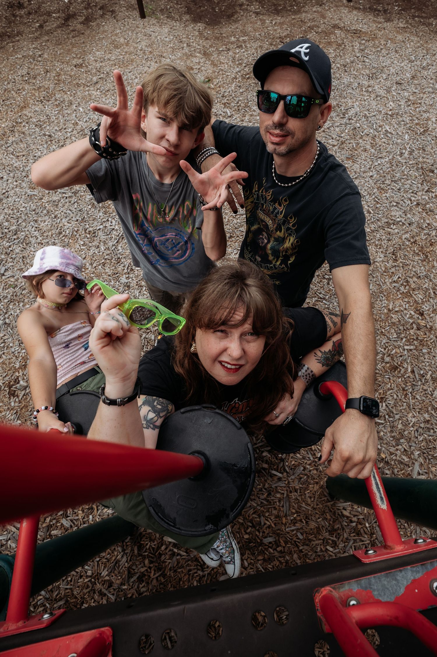 Four people pose playfully on red playground equipment over wood chips, one boy flashing hand gestures and a woman holding green glasses.