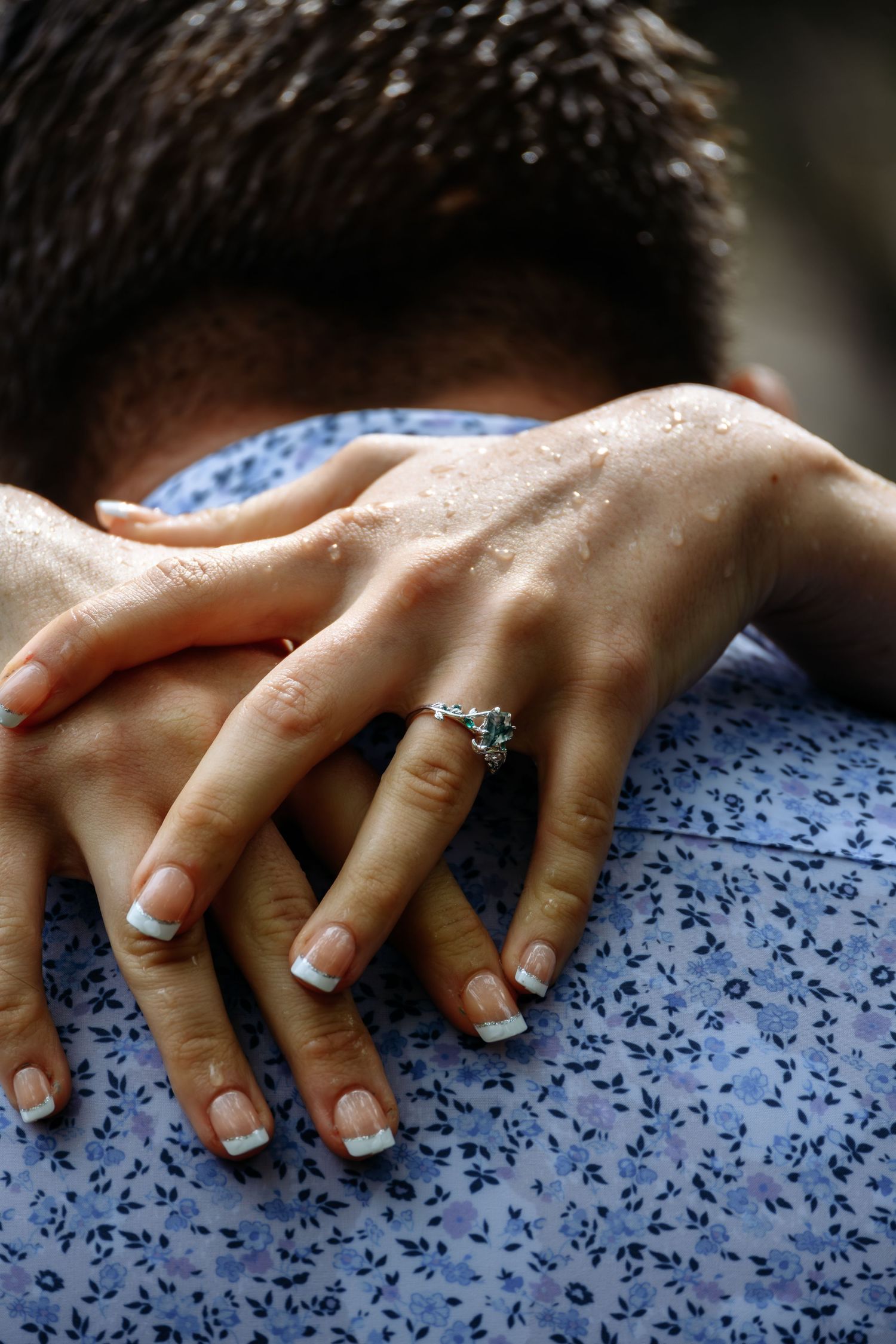 Close-up of womans hands with diamond engagement ring clasped on mans wet blue shirt.