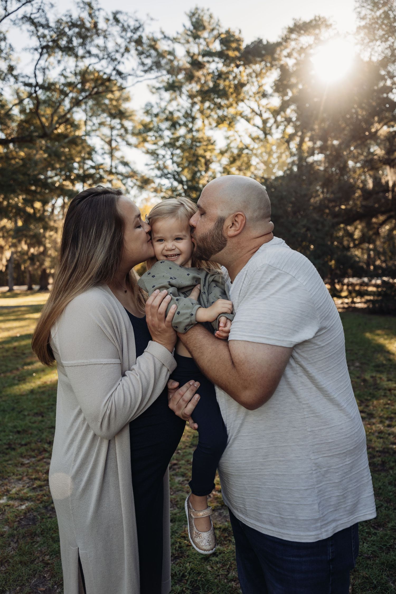 Parents kissing their smiling toddler on the cheeks outdoors in a sunlit park with lush green trees.