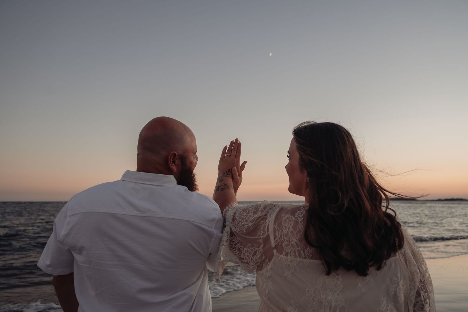 Couple standing on a beach at sunset, facing the ocean as the woman raises her hand toward a pale moon in the pink and blue sky.