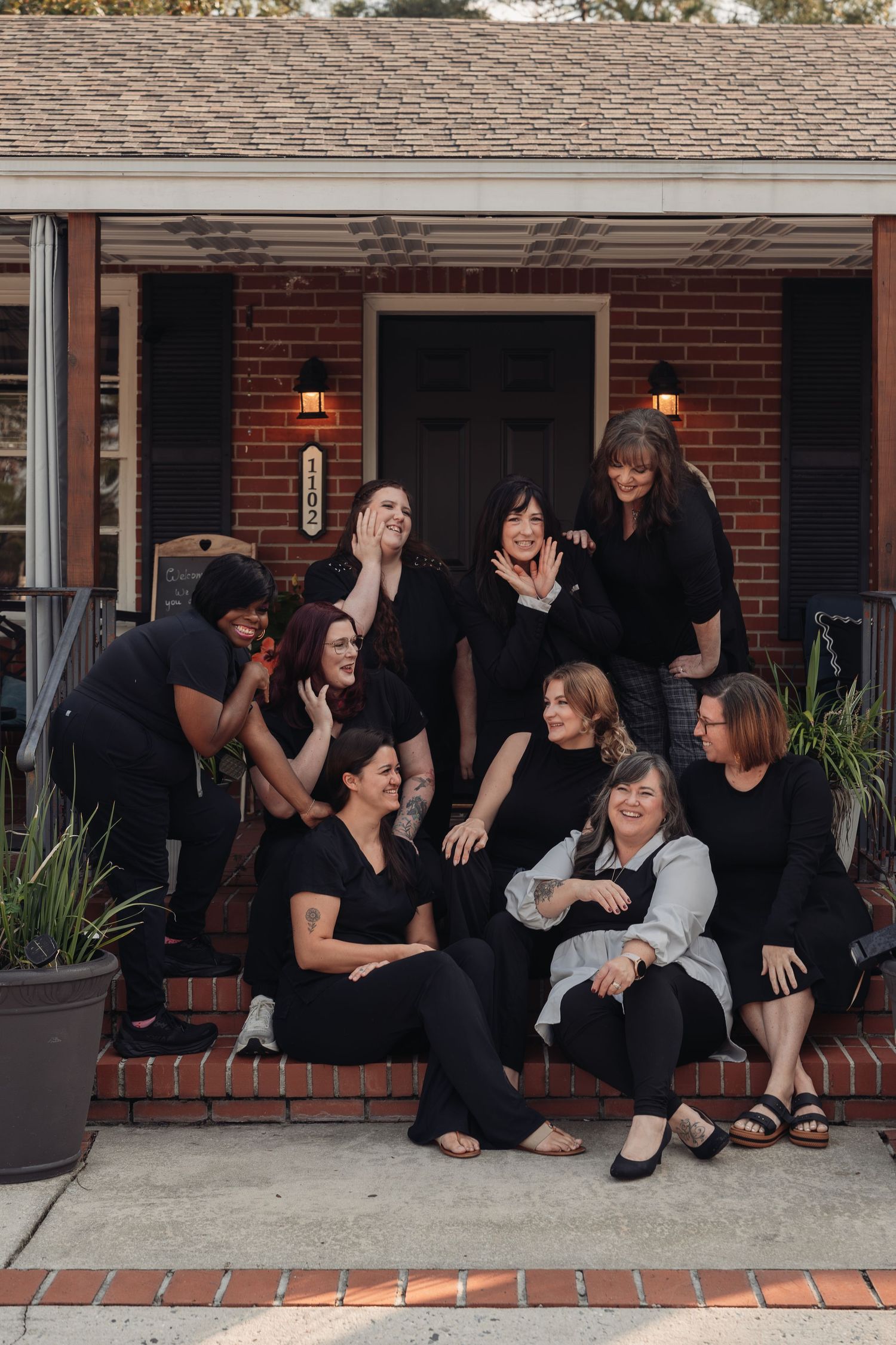 A group of nine women in black outfits laugh and pose together on the brick front steps of a house with a dark door and lit lanterns.
