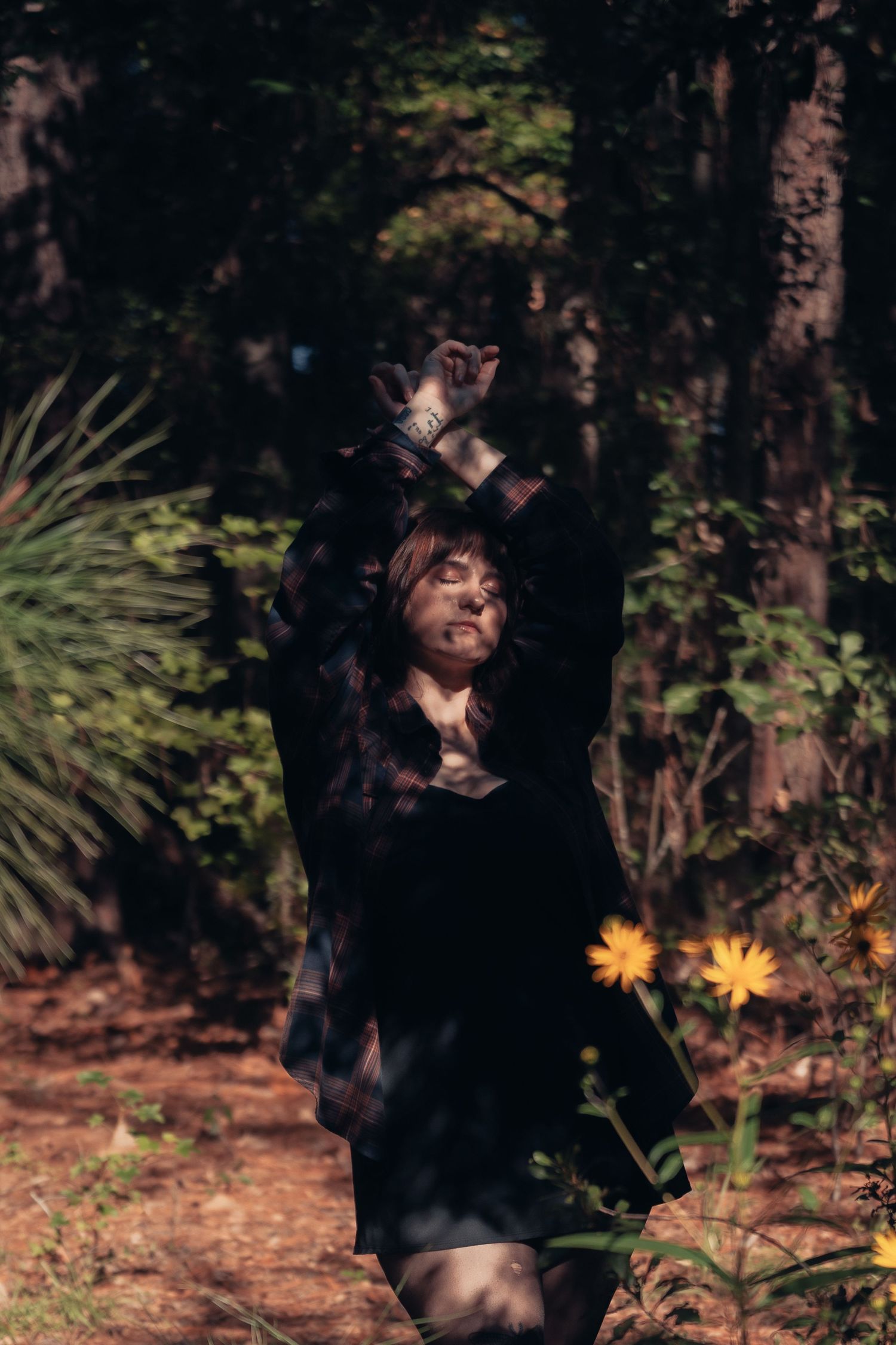 A woman in a dark dress stands among forest trees and yellow wildflowers, arms raised, bathed in dappled sunlight.