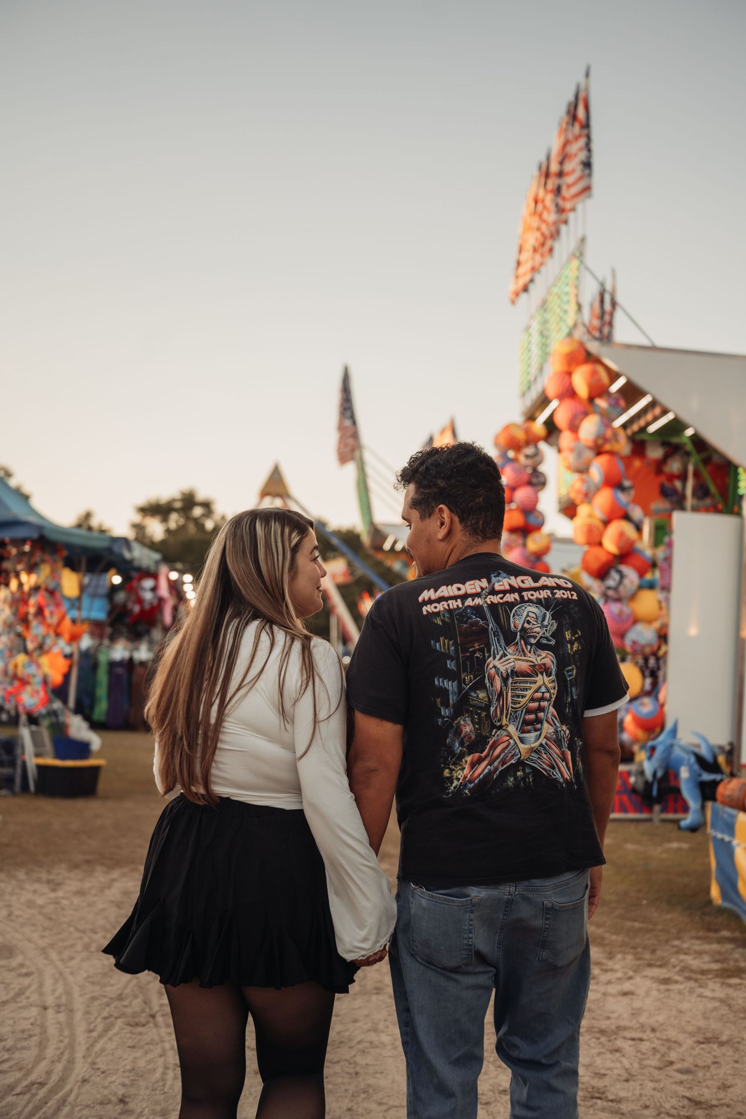 Couple holding hands and walking away from the camera toward colorful carnival game booths and flags at a fairground at sunset.