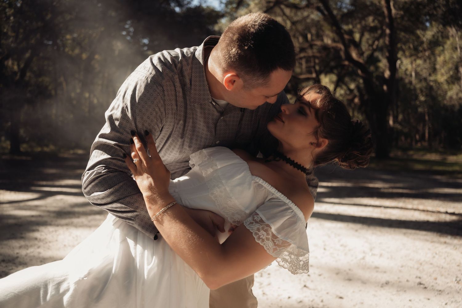 Man dipping woman dramatically in a sun-dappled forest, woman in white off-shoulder lace wedding dress, romantic outdoor shoot.