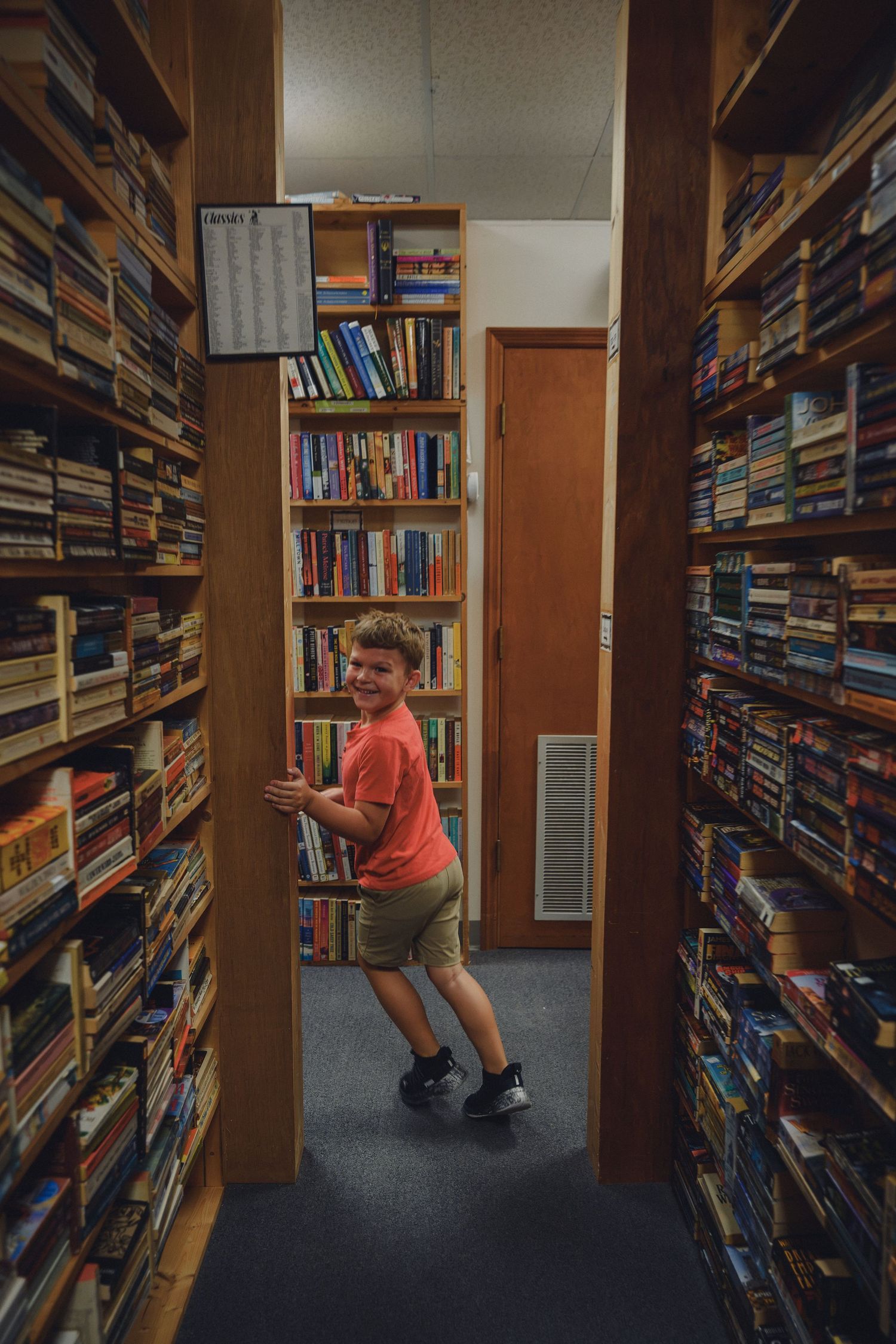 A smiling boy in an orange shirt explores narrow aisles lined with books in a cozy used bookstore.
