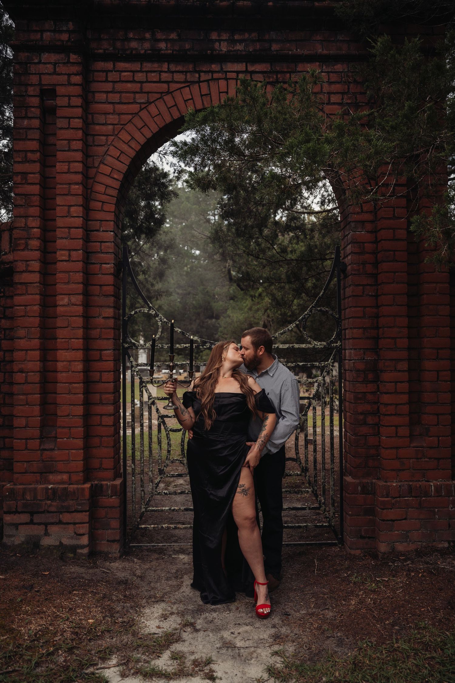 Couple kissing in a romantic brick archway with ornate iron gates, woman in black gown with red heels, moody outdoor setting.