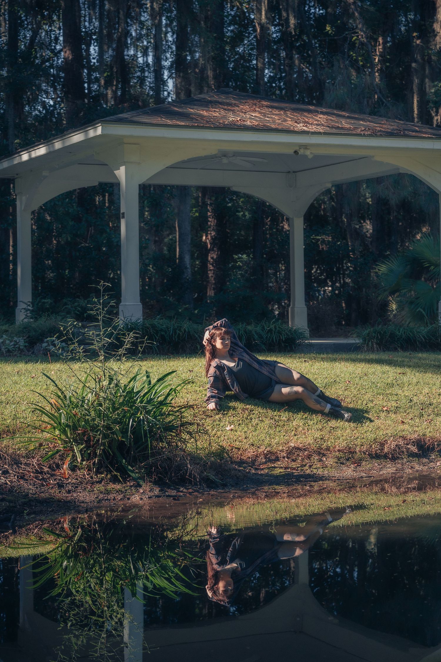 Woman in black dress reclining by a pond near a white gazebo, with her reflection visible in the still water.