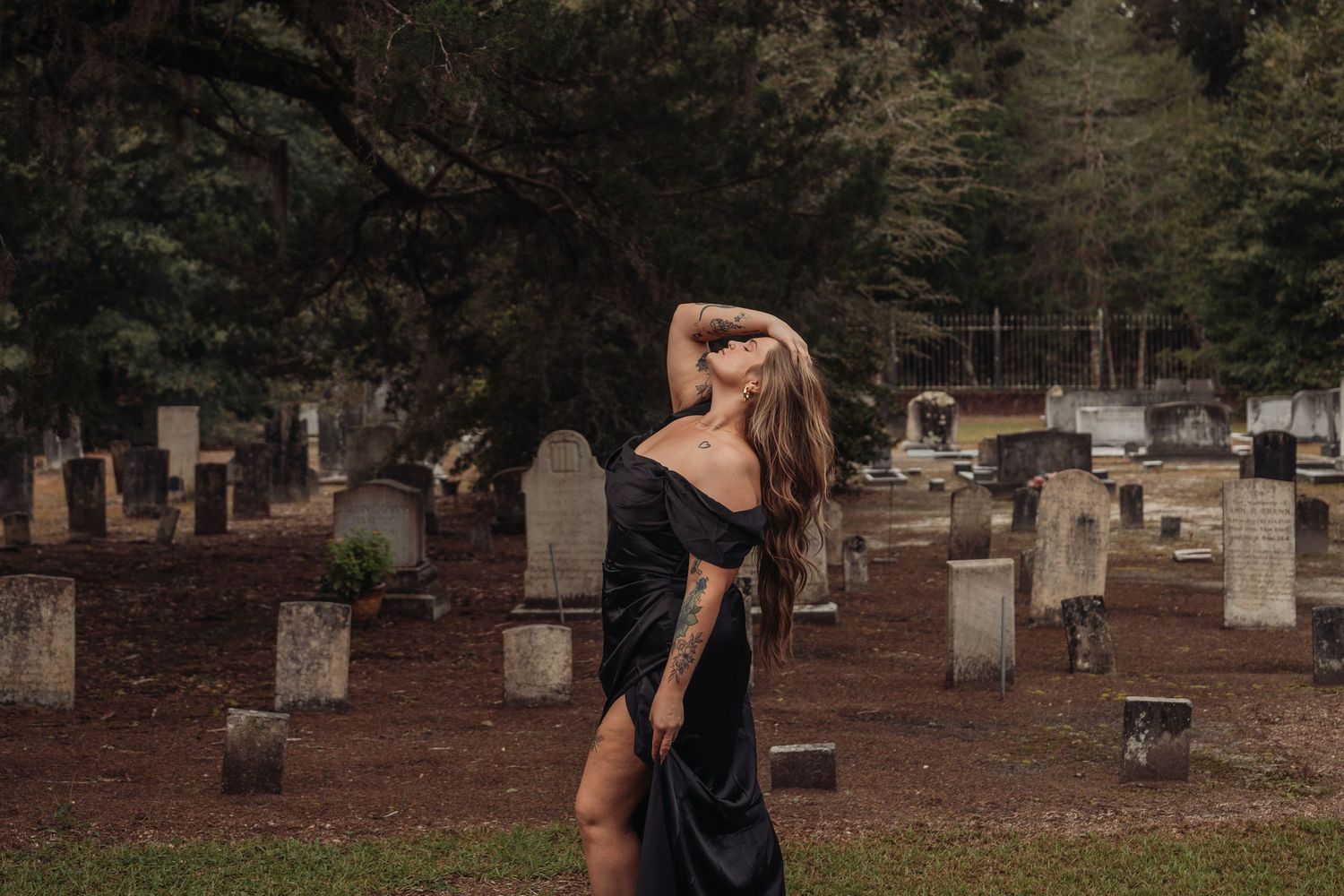 Woman in black dress posing among gravestones in a cemetery, with trees in the background.