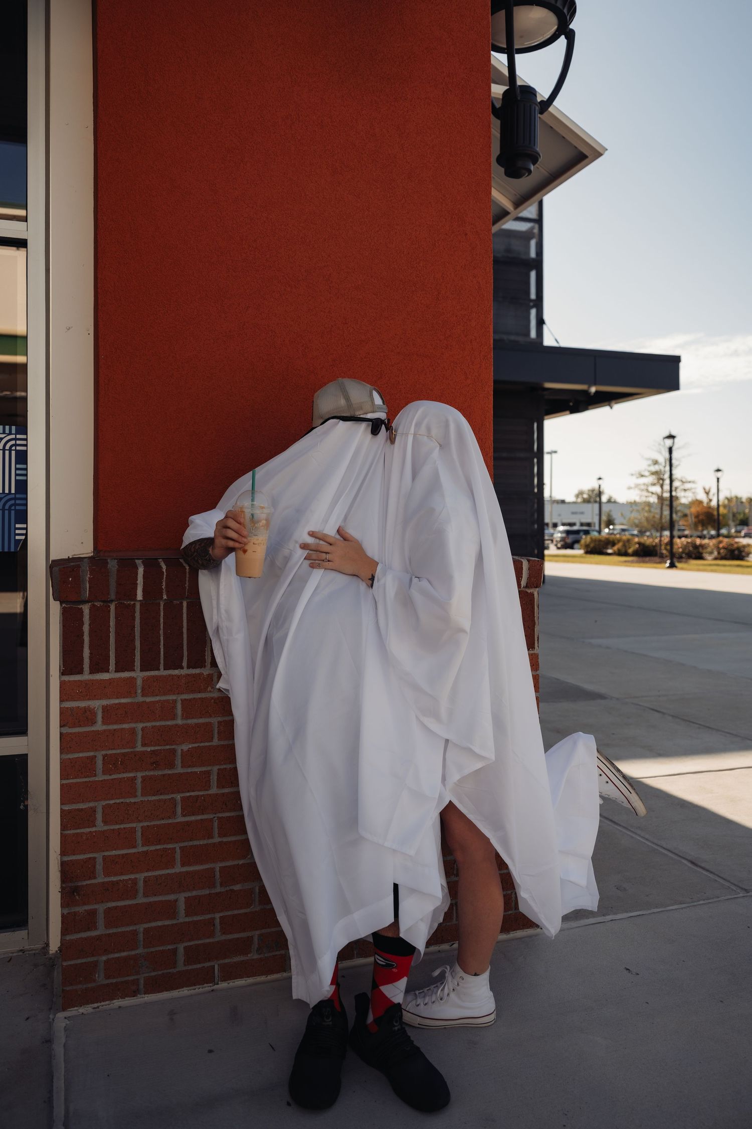 Two people dressed as ghosts in white sheets embrace against a red brick wall outside a store.