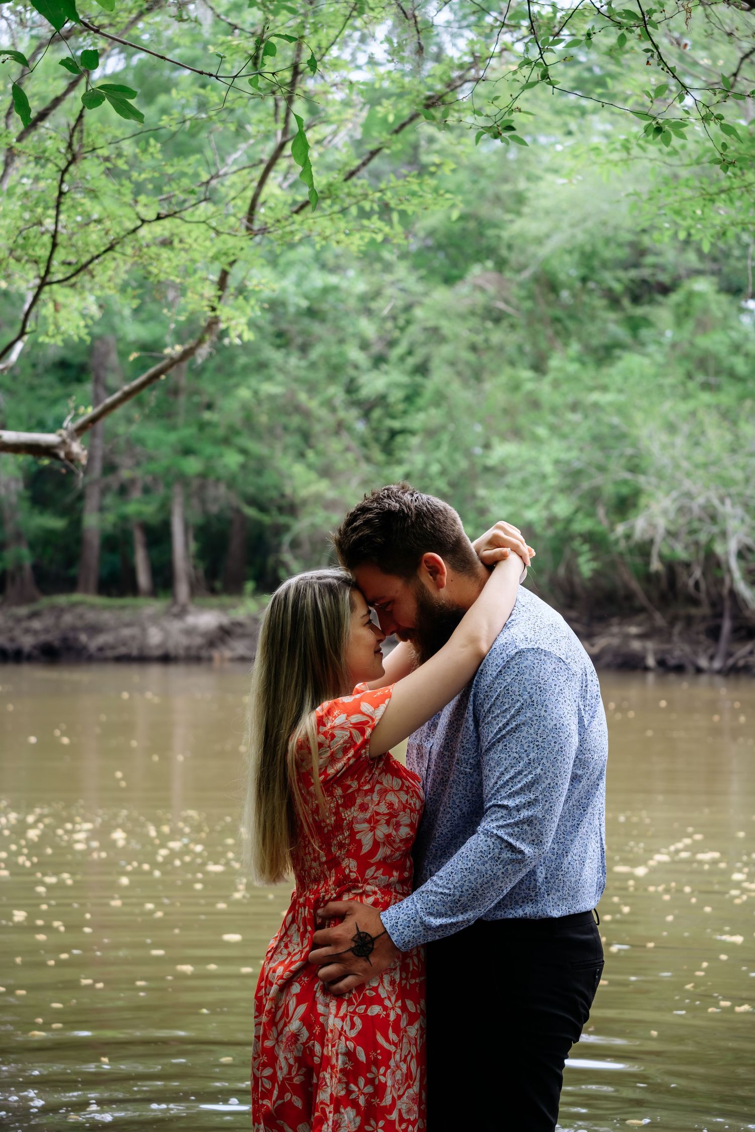 Couple shares a tender almost-kiss while standing in a shallow river surrounded by lush green forest.