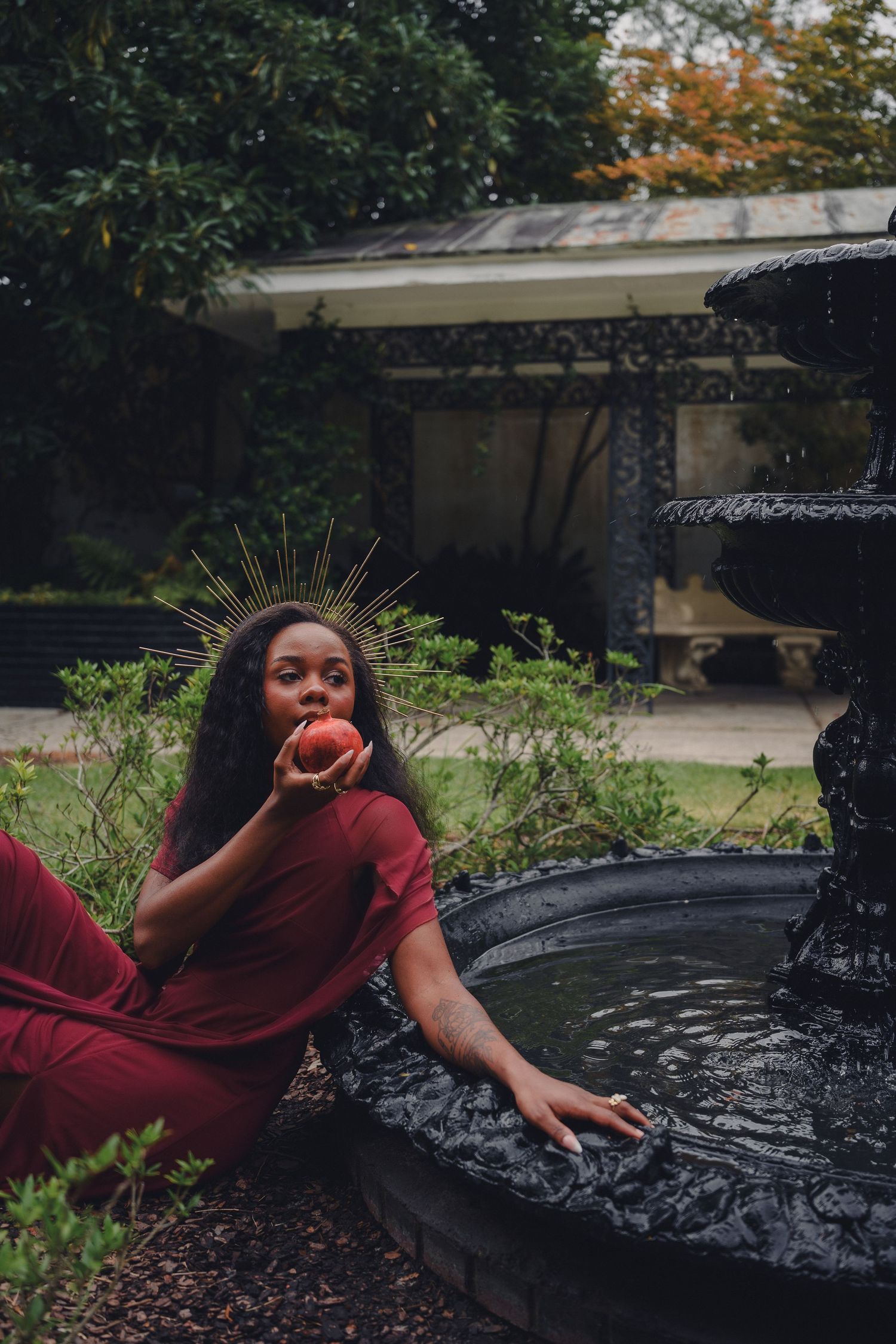 A woman in a red dress wears a gold halo crown, holding an apple beside a dark ornate fountain in a lush garden.