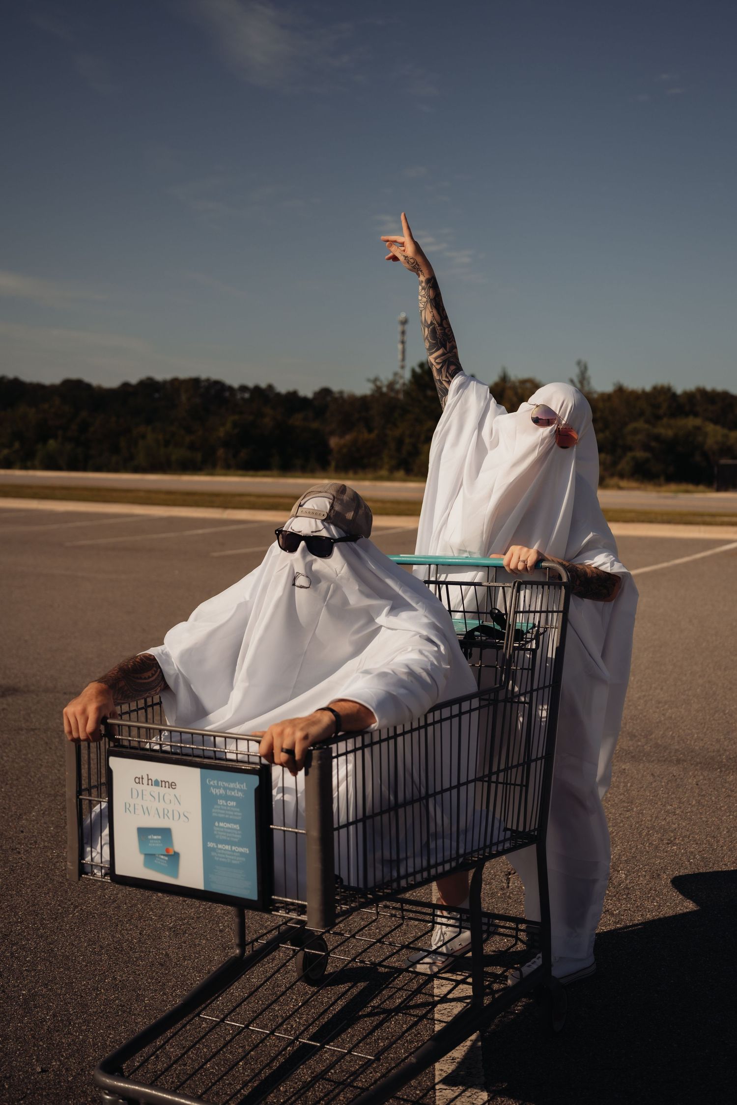 Two people dressed as ghosts pose in a shopping cart in a parking lot, one pointing upward.